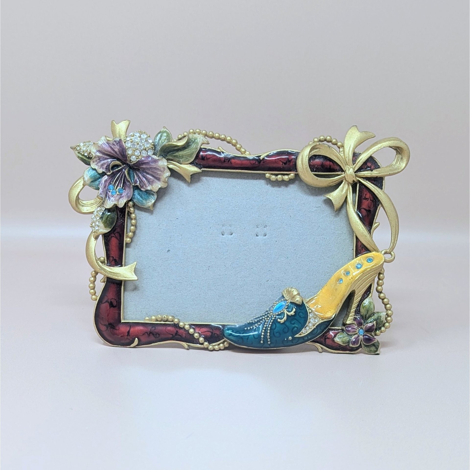 A horizontal photo frame with a deep red enamel border. Ornate gold-tone decorations include a crystal-studded purple hibiscus-style flower on the top left and a fashionable stiletto shoe on the bottom right.