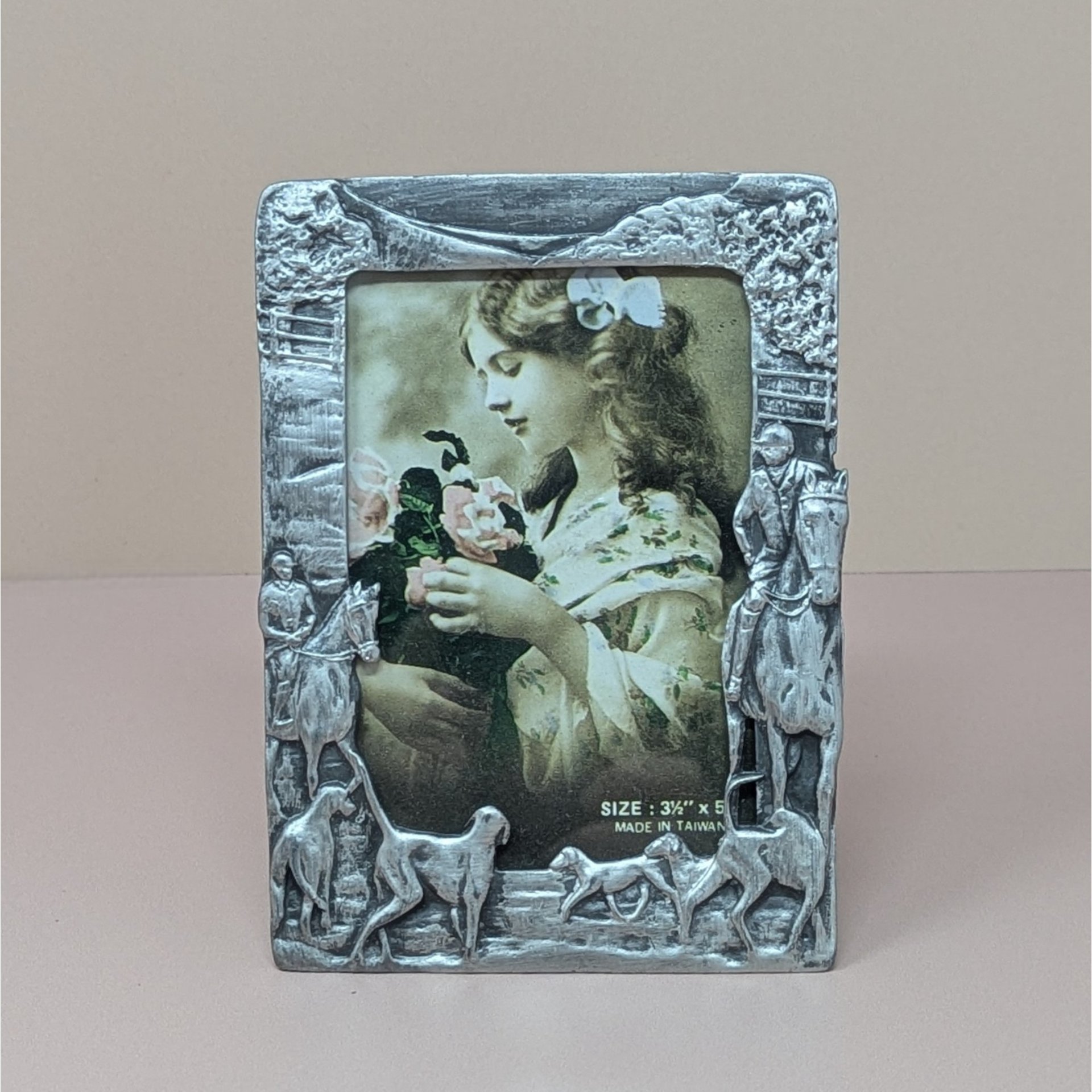 A vertical cast-metal picture frame with a weathered silver finish. Embossed hunters on horses and a pack of running hounds frame a portrait of a young girl with flowers.