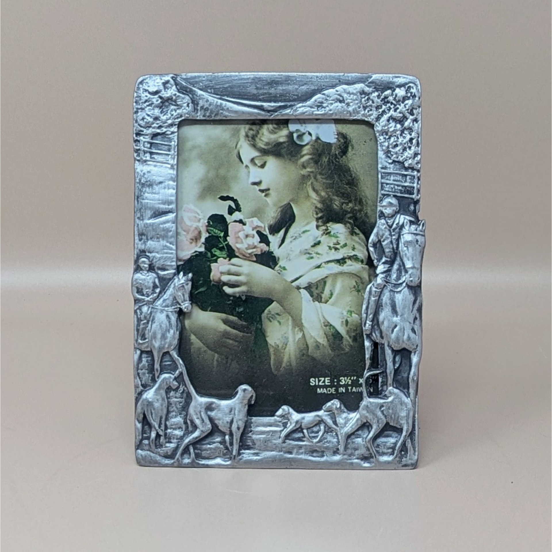 A vertical cast-metal picture frame with a weathered silver finish. Embossed hunters on horses and a pack of running hounds frame a portrait of a young girl with flowers.