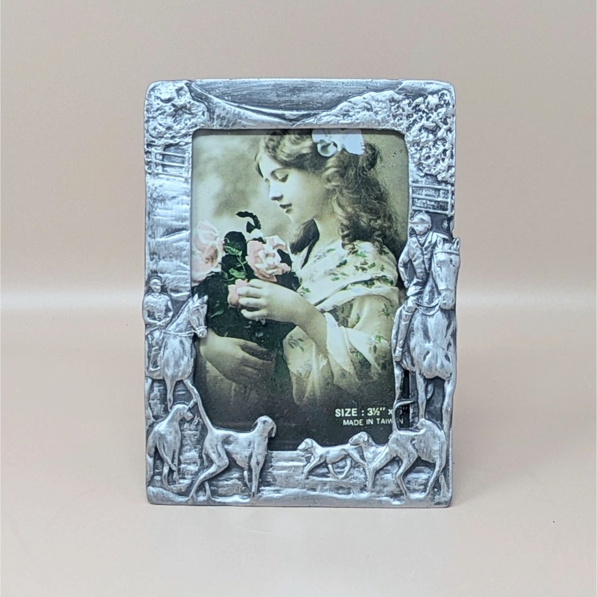 A vertical cast-metal picture frame with a weathered silver finish. Embossed hunters on horses and a pack of running hounds frame a portrait of a young girl with flowers.