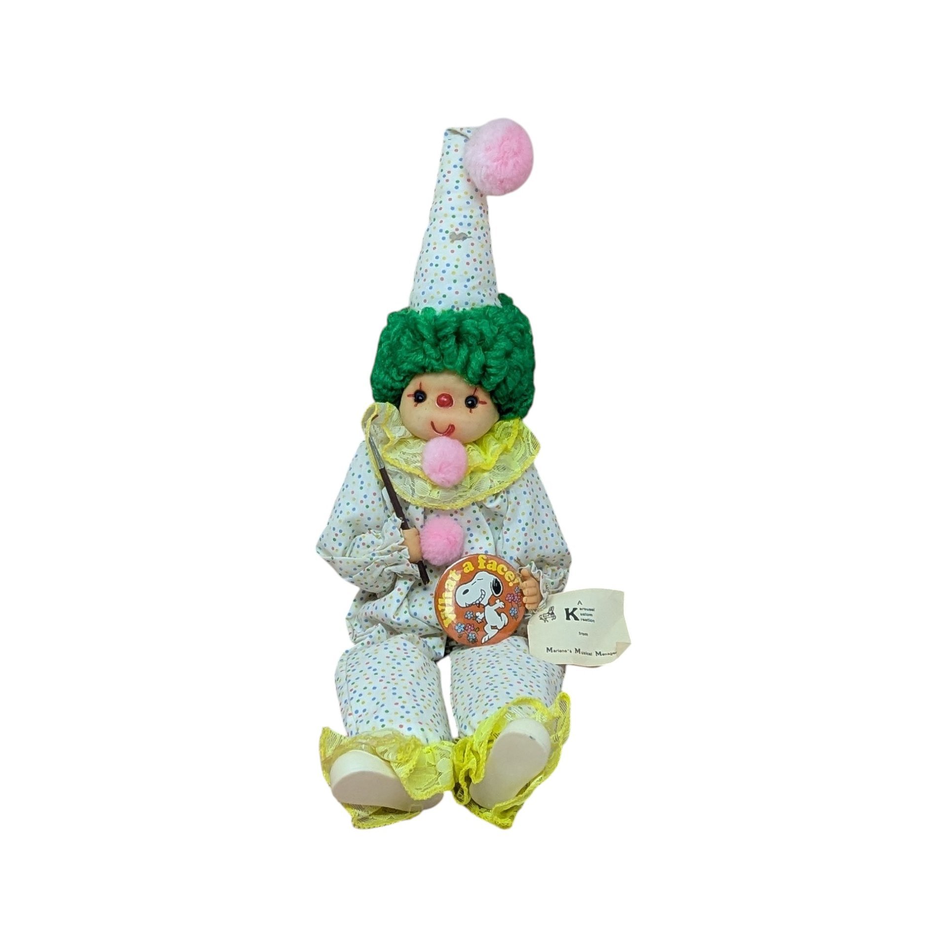 A playful clown doll sitting upright, wearing a white outfit with tiny colorful dots and yellow ruffles. It has green curly yarn hair and three pink pom-poms down its front, holding a small paint brush.