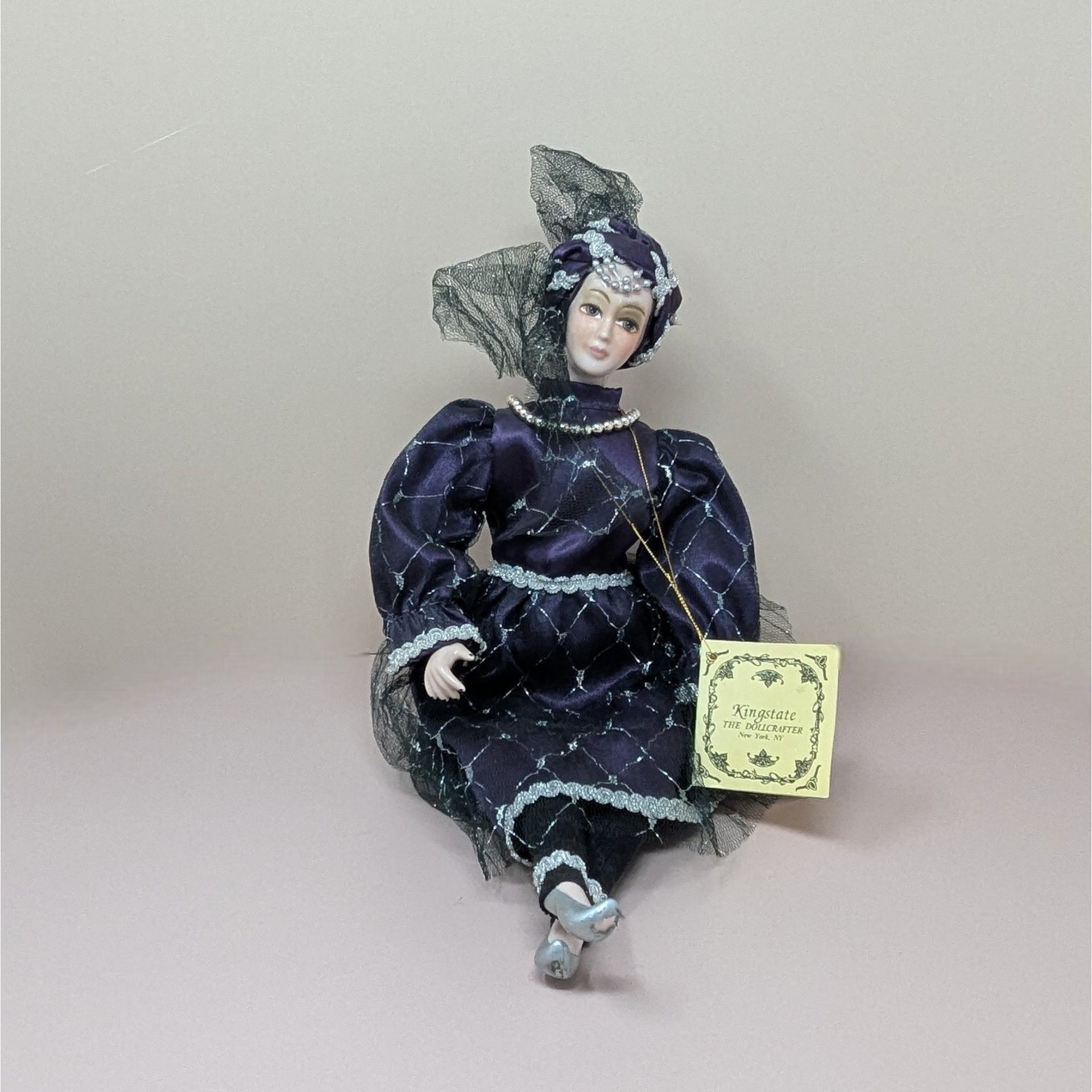 A vintage Kingstate porcelain harlequin doll in a deep purple satin dress with silver lattice patterns and lace trim. She features hand-painted facial features and an ornate turban with a black mesh veil.