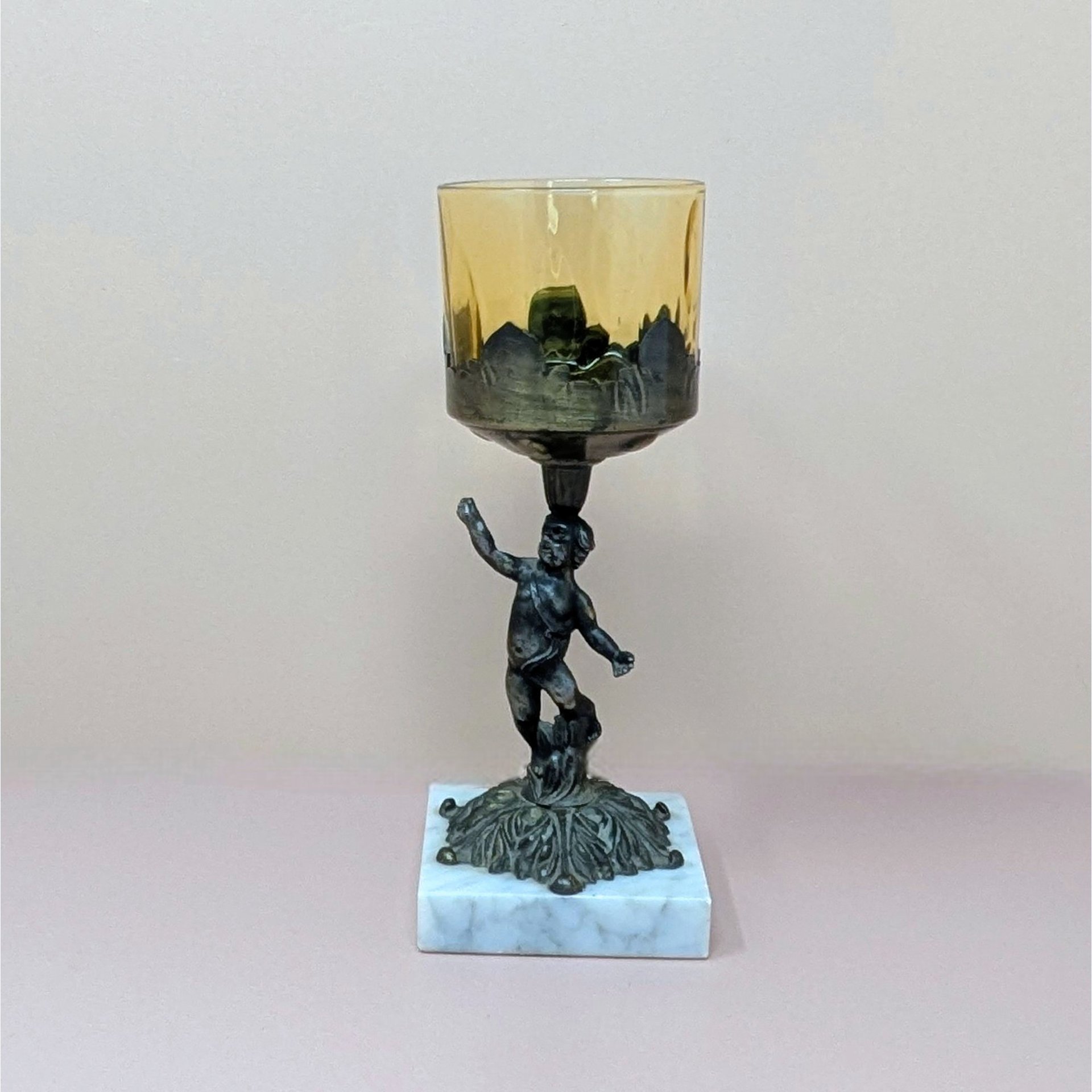 A 9-inch vintage candle holder featuring a dark cast metal cherub figure standing on a white marble square base, supporting a translucent amber glass cup.