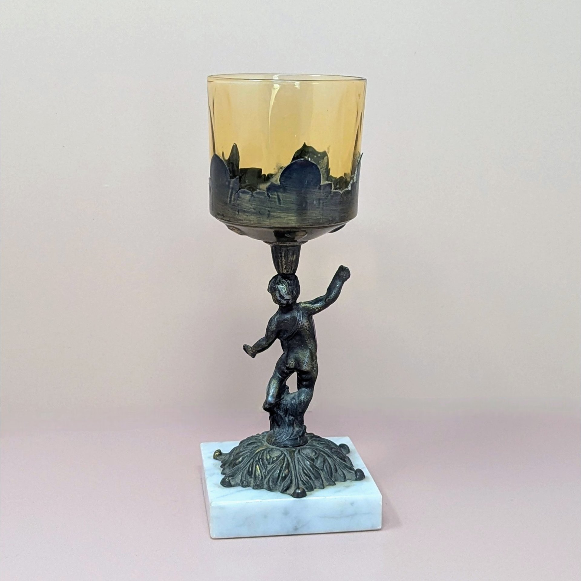 A 9-inch vintage candle holder featuring a dark cast metal cherub figure standing on a white marble square base, supporting a translucent amber glass cup.