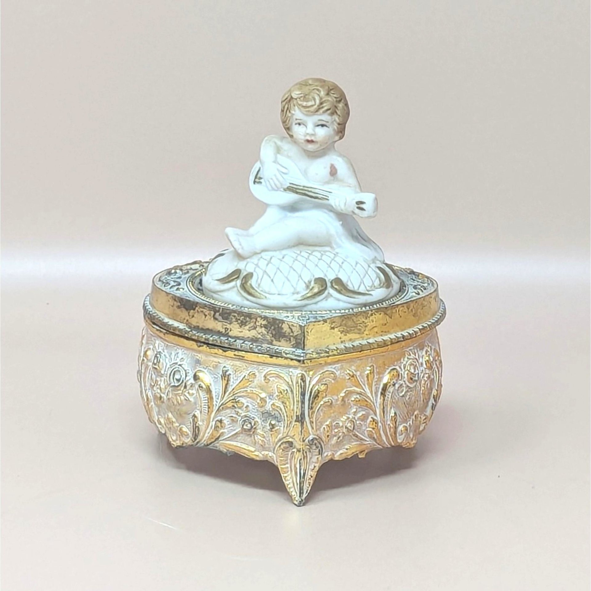 A vintage footed trinket box with a highly detailed gold-toned floral base and a white bisque-style lid featuring a cherub playing a stringed instrument.