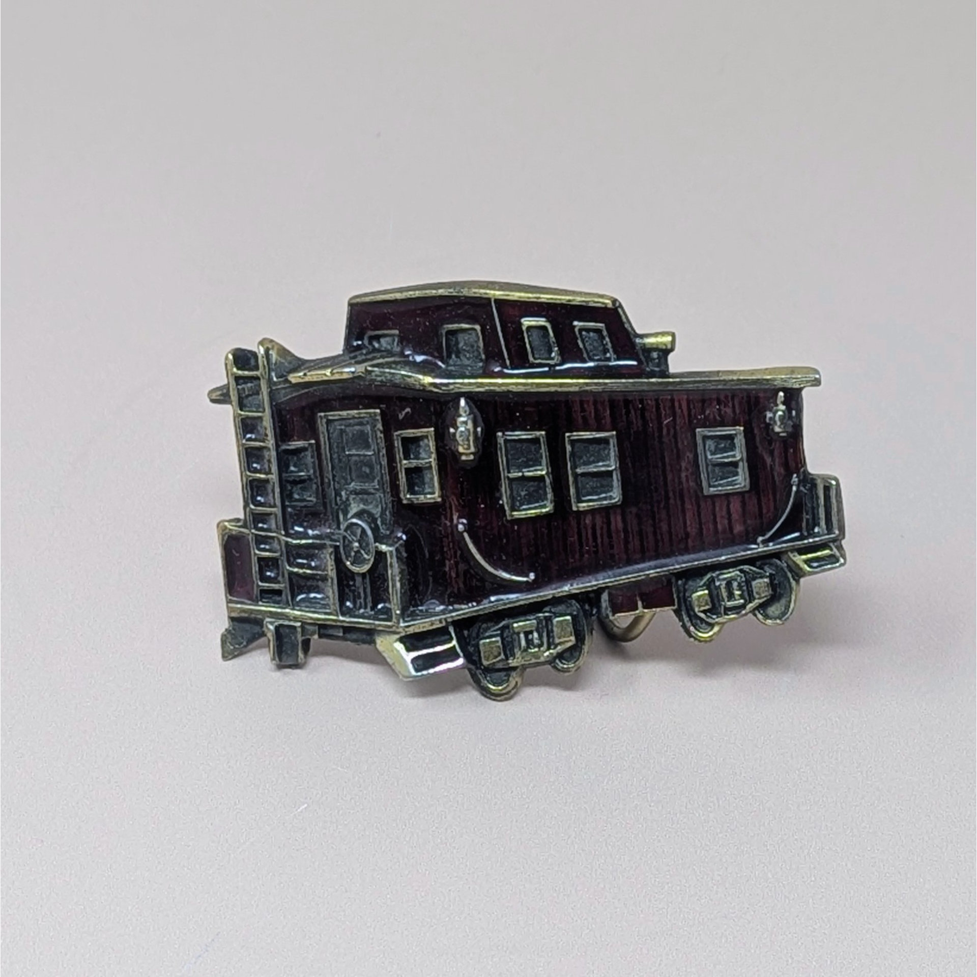 A vintage 1976 belt buckle shaped like a train caboose with deep red enamel and brass-toned metal accents on a neutral background.