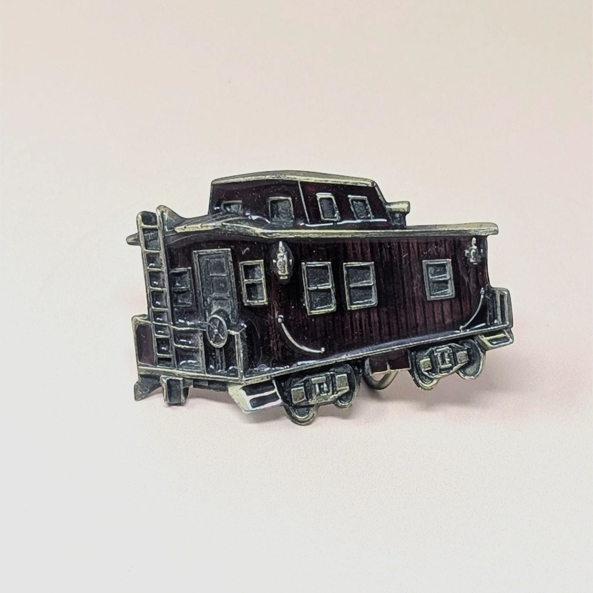 A vintage 1976 belt buckle shaped like a train caboose with deep red enamel and brass-toned metal accents on a neutral background.