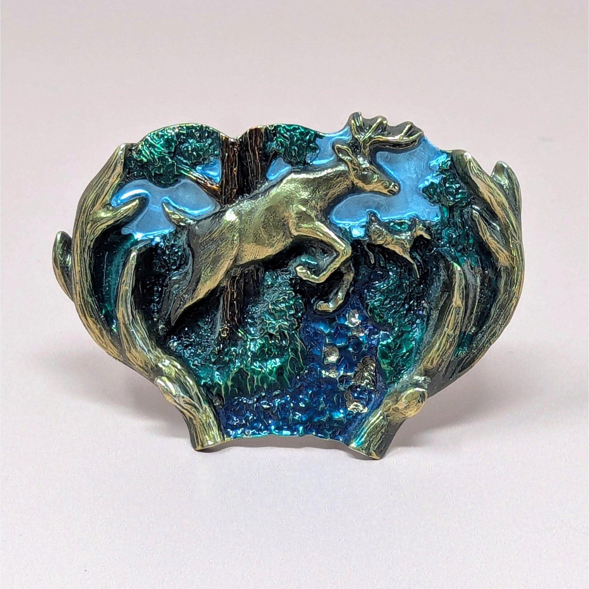 A vintage 1982 belt buckle featuring a buck and doe leaping through a green forest with a blue sky, framed by a brass-toned antler border.