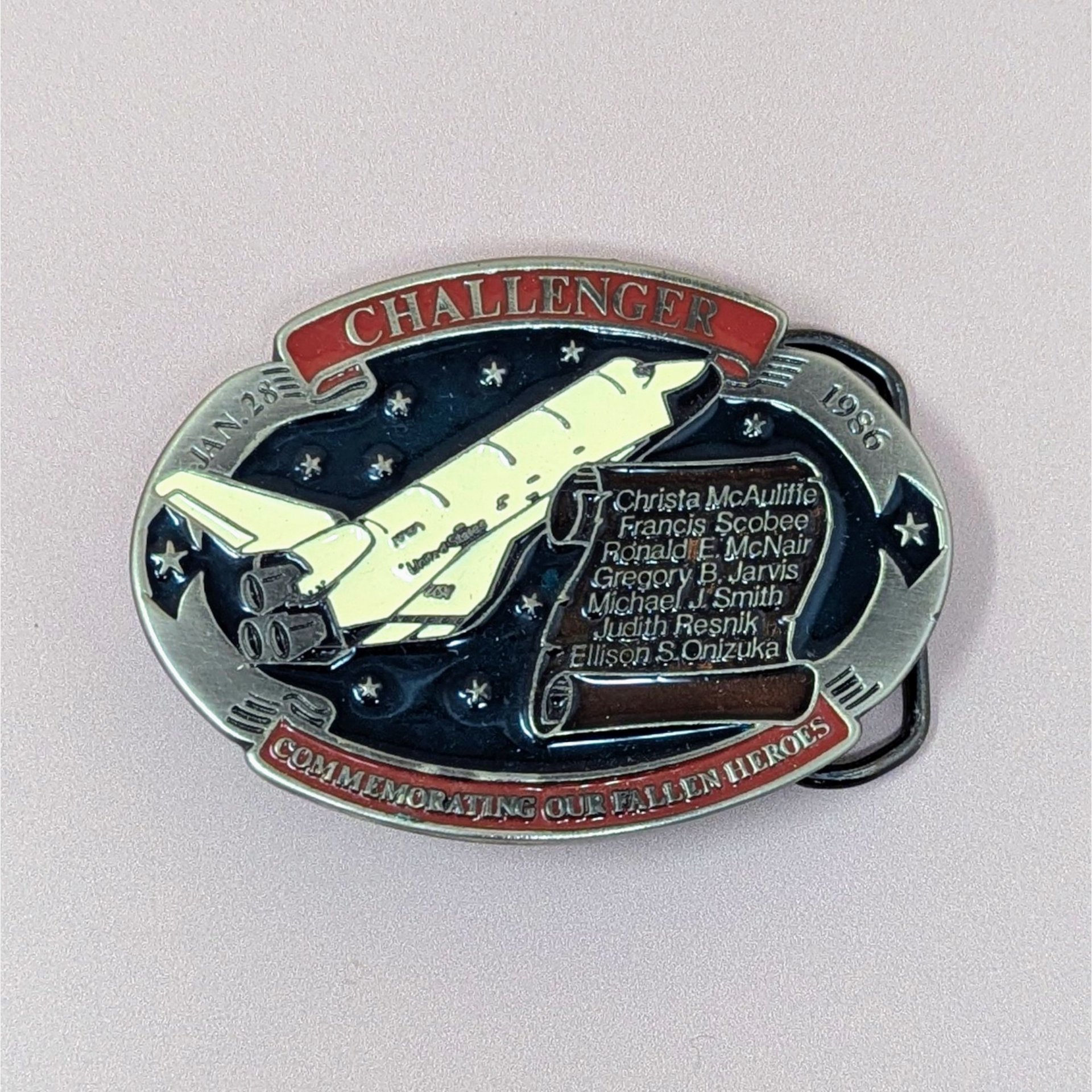 This Challenger Belt Buckle features an oval silhouette and a metallic texture, offering a reverent aesthetic for a curated study.   This finely detailed commemorative piece was produced in 1986 to honor the crew of the Space Shuttle Challenger mission STS-51-L. The buckle showcases a high-relief white shuttle ascending against a deep black enamel sky dotted with silver stars. A red scroll at the top boldly displays the Challenger name, while a matching banner along the bottom edge reads "Commemorating Our 
