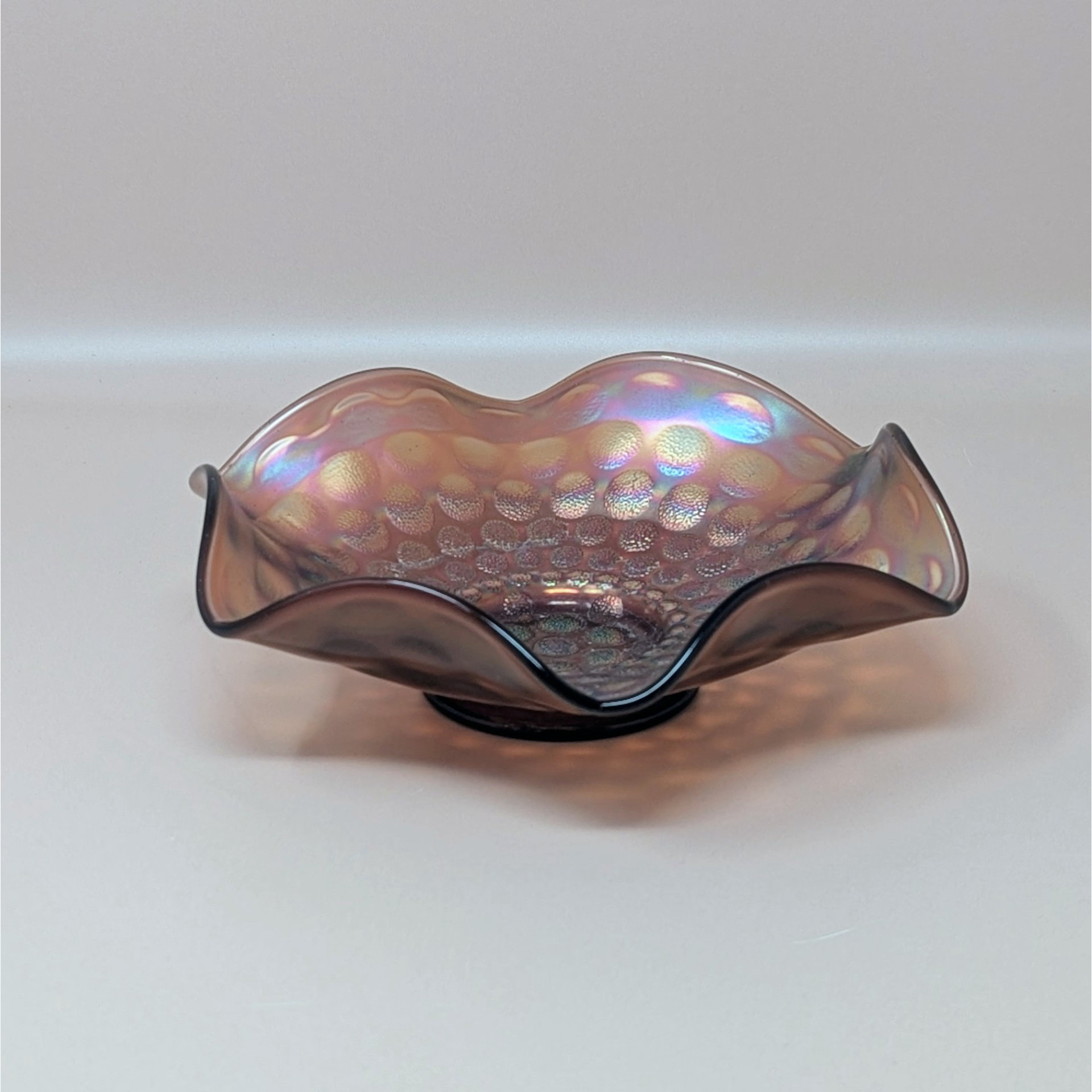A vintage Fenton marigold carnival glass bowl with a ruffled edge and a dimpled coin dot pattern, showing iridescent pink and gold tones.