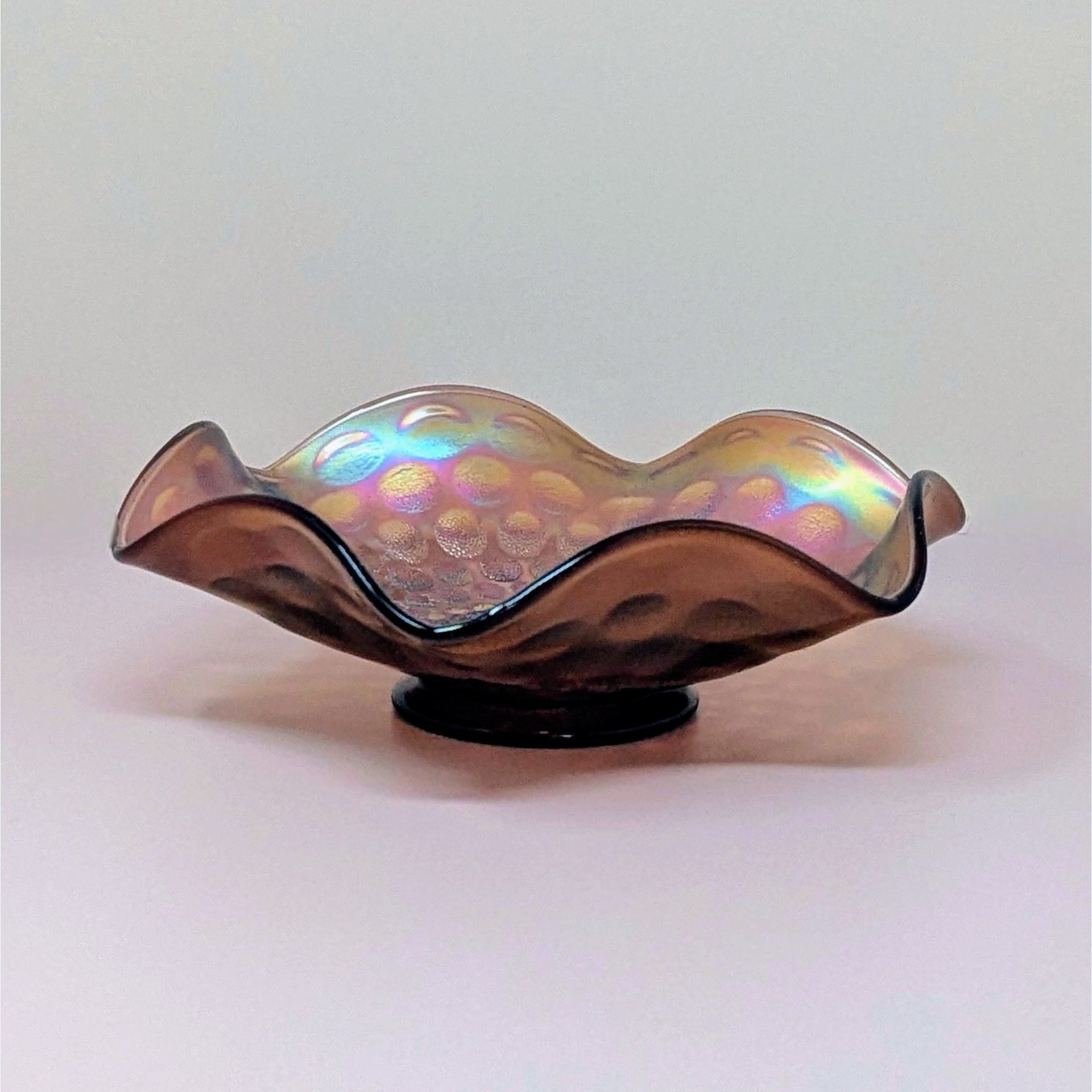 A vintage Fenton marigold carnival glass bowl with a ruffled edge and a dimpled coin dot pattern, showing iridescent pink and gold tones.