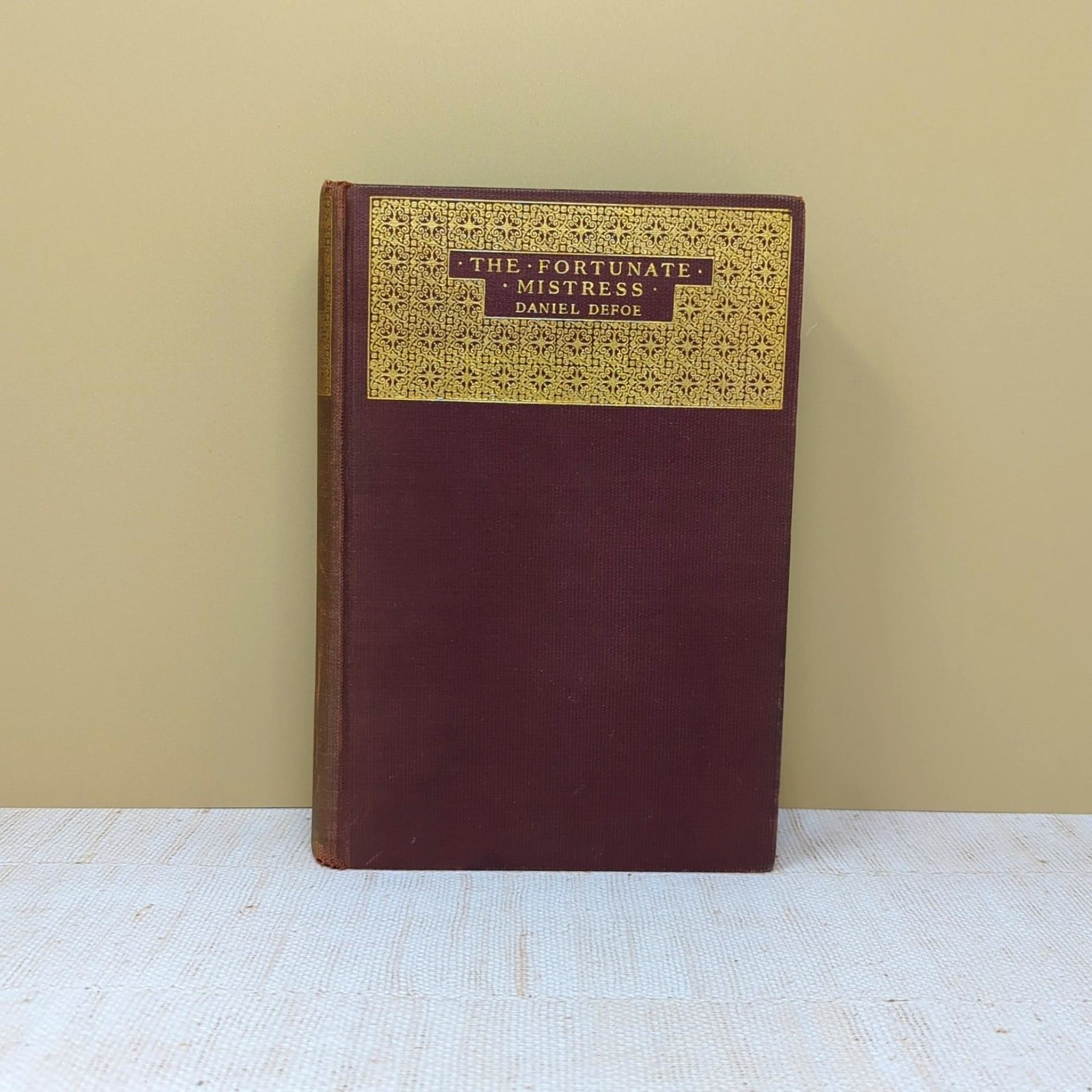 A plum-colored hardcover book with a large gold-stamped ornate panel on the front cover titled The Fortunate Mistress by Daniel Defoe against a neutral background.
