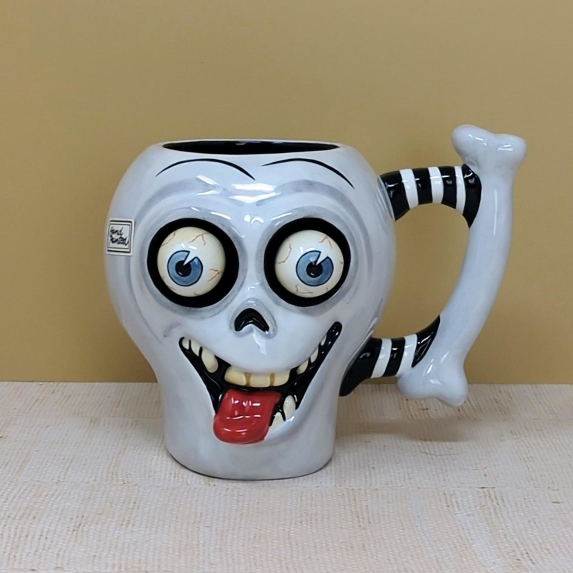 A whimsical white ceramic skull mug with large bulging eyes, a protruding red tongue, and a black-and-white striped bone handle against a neutral background.