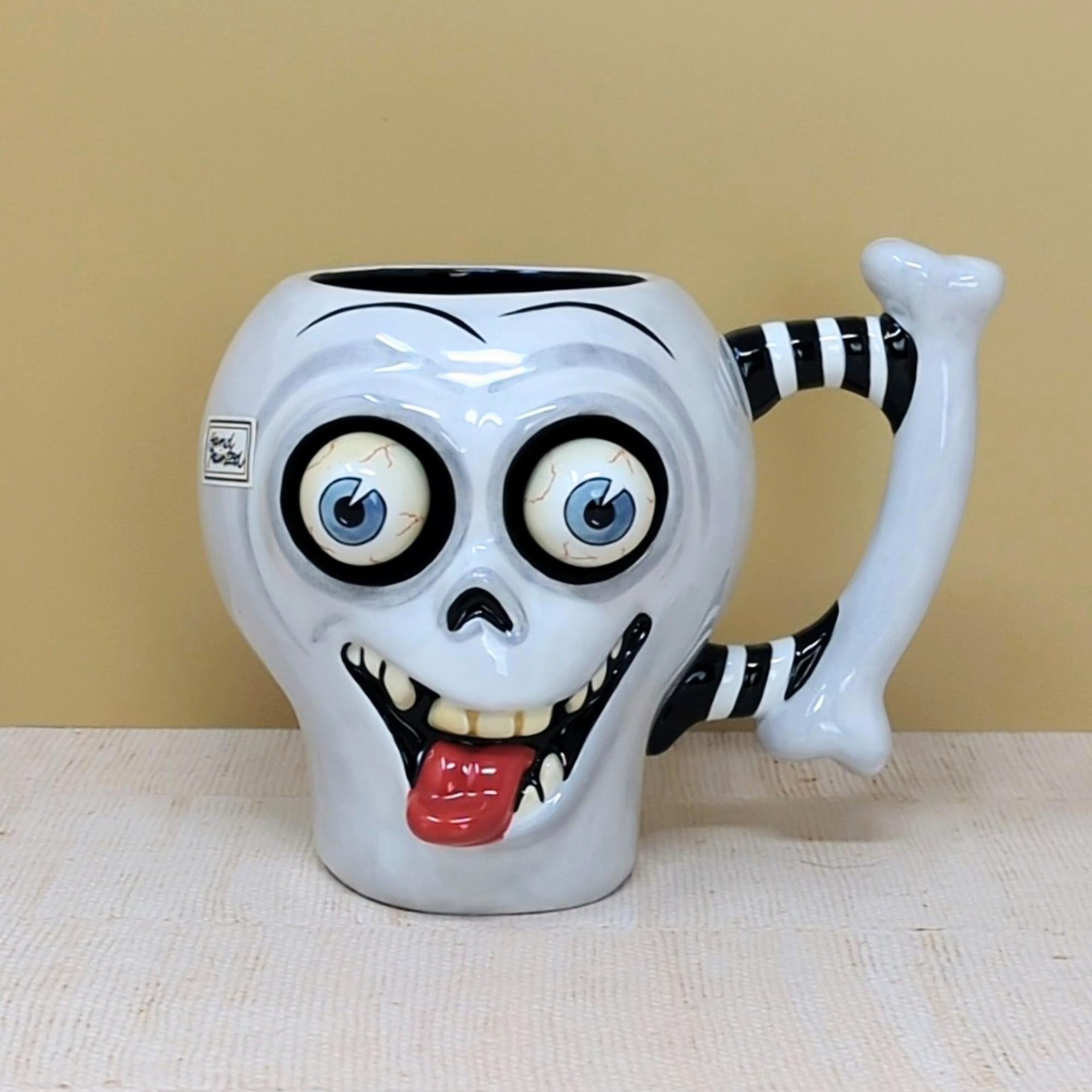 A whimsical white ceramic skull mug with large bulging eyes, a protruding red tongue, and a black-and-white striped bone handle against a neutral background.