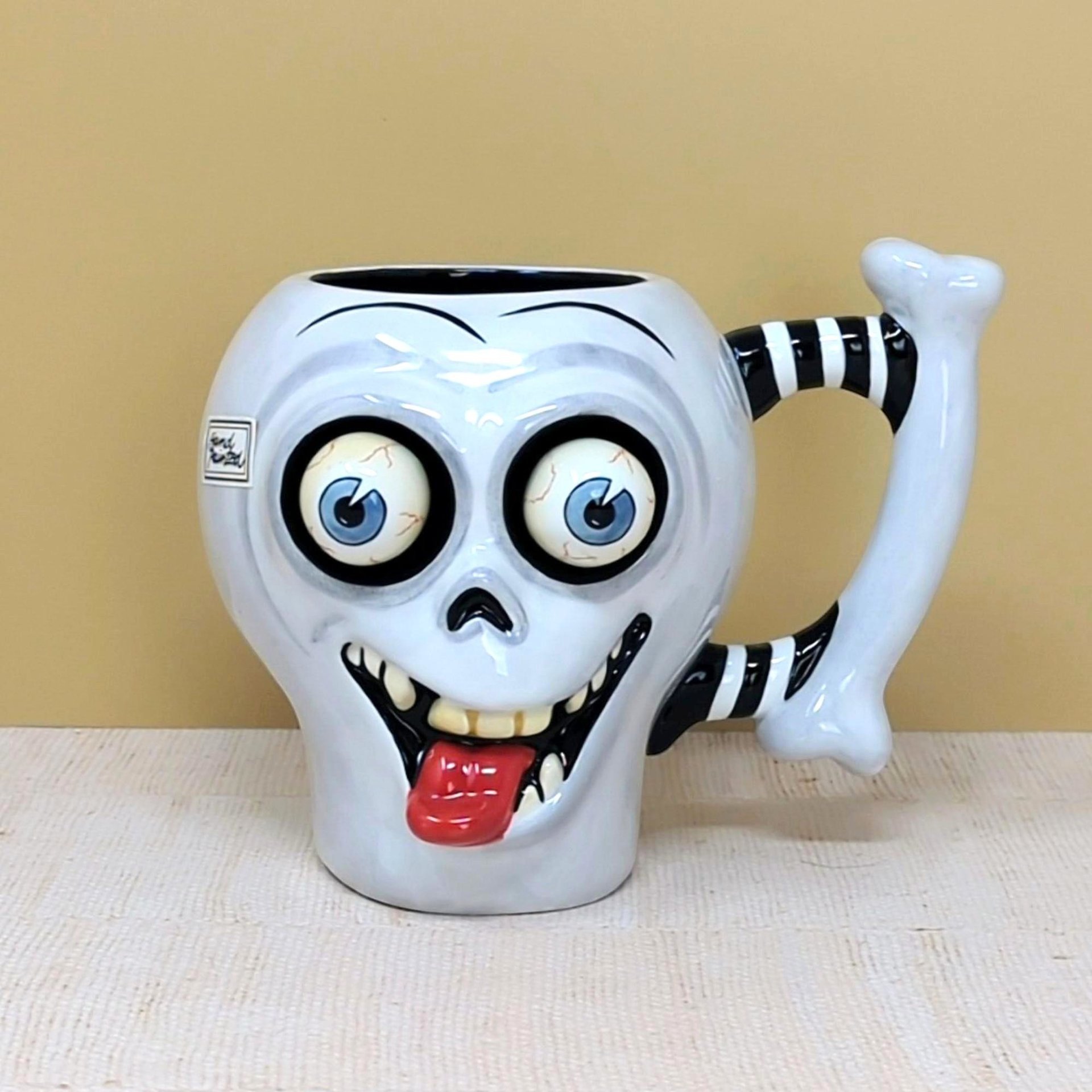 A whimsical white ceramic skull mug with large bulging eyes, a protruding red tongue, and a black-and-white striped bone handle against a neutral background.