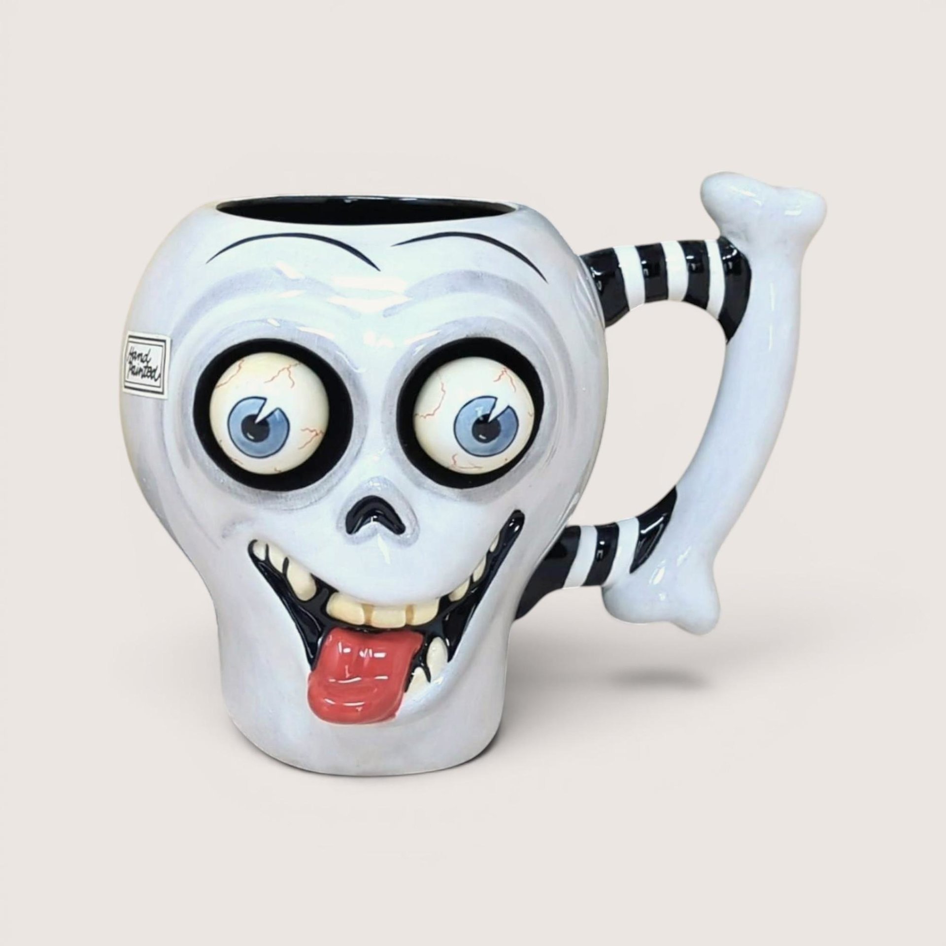 A whimsical white ceramic skull mug with large bulging eyes, a protruding red tongue, and a black-and-white striped bone handle against a neutral background.