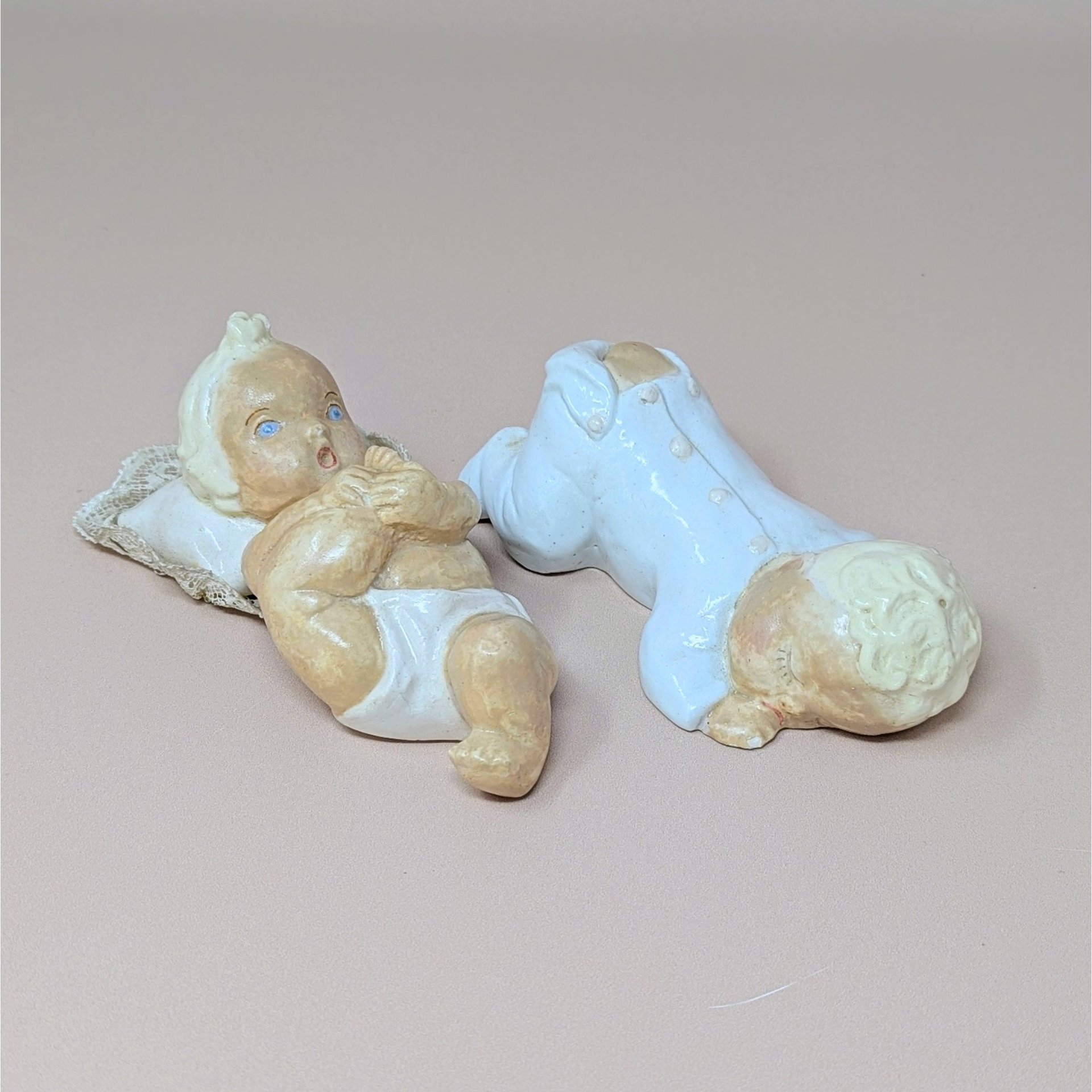 A pair of hand-painted chalkware baby figurines, one reclining on a pillow and the other in a white sleeper, against a neutral background.