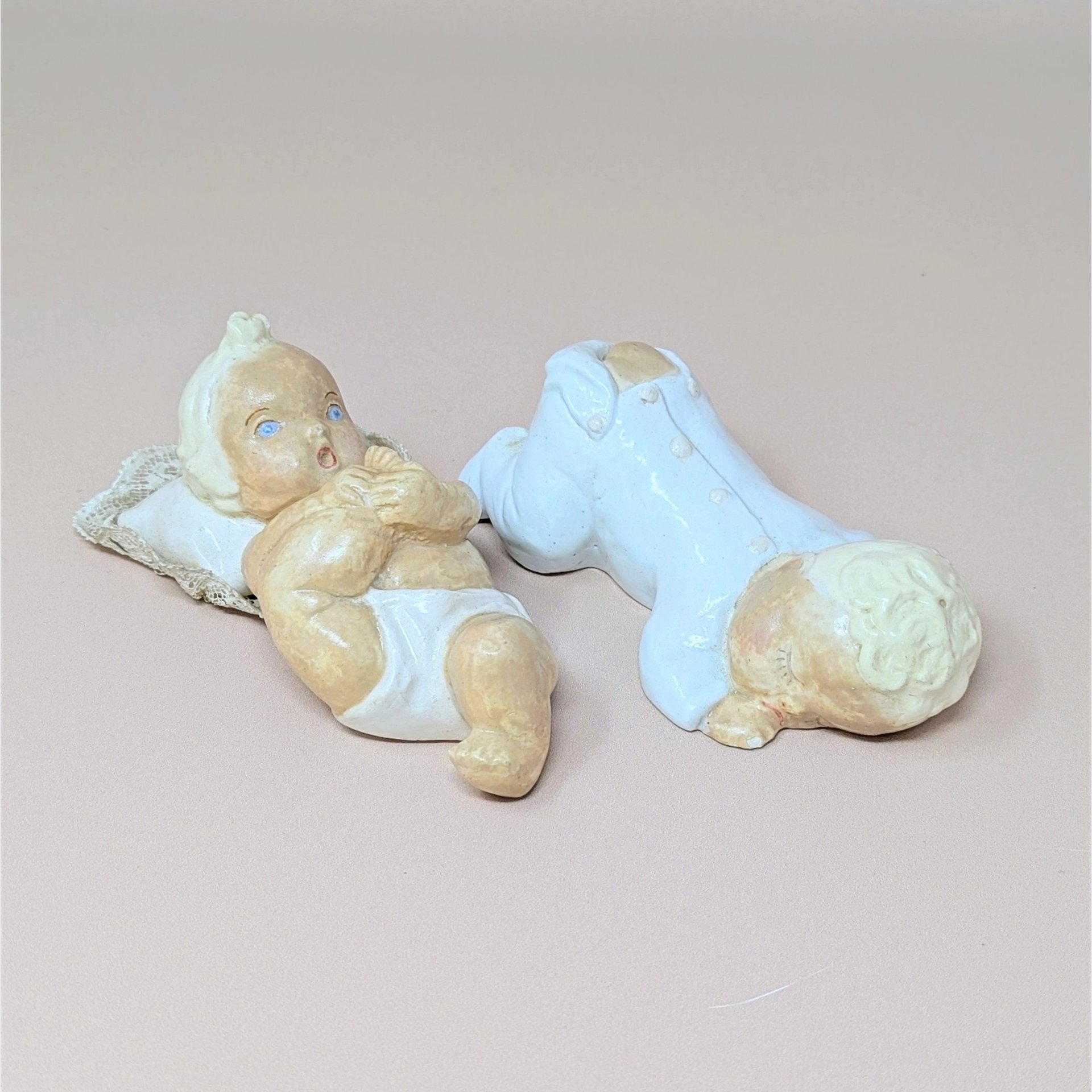A pair of hand-painted chalkware baby figurines, one reclining on a pillow and the other in a white sleeper, against a neutral background.