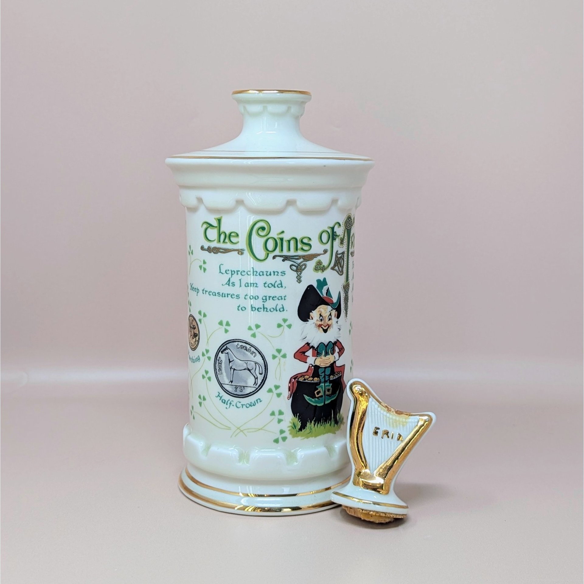 A tall white porcelain decanter featuring colorful Irish coin graphics, a leprechaun illustration, and a gold-trimmed harp stopper against a neutral background.