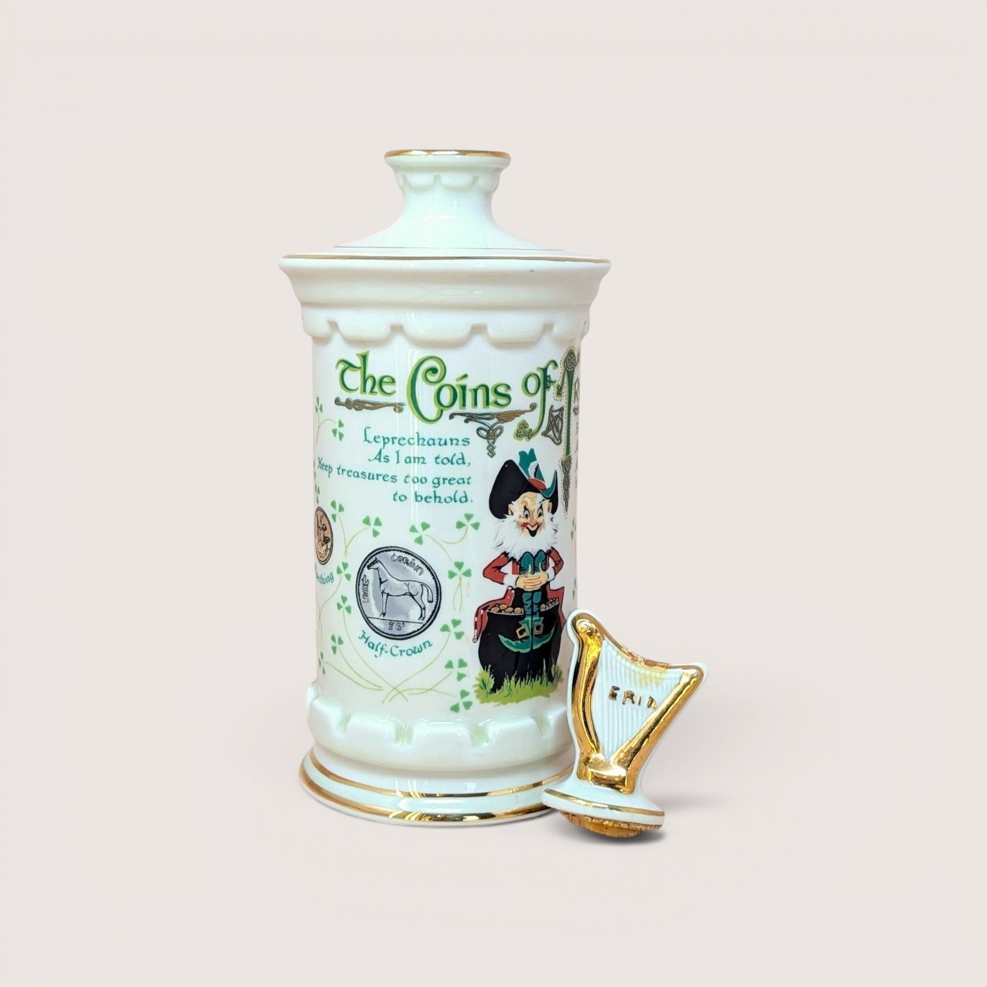 A tall white porcelain decanter featuring colorful Irish coin graphics, a leprechaun illustration, and a gold-trimmed harp stopper against a neutral background.