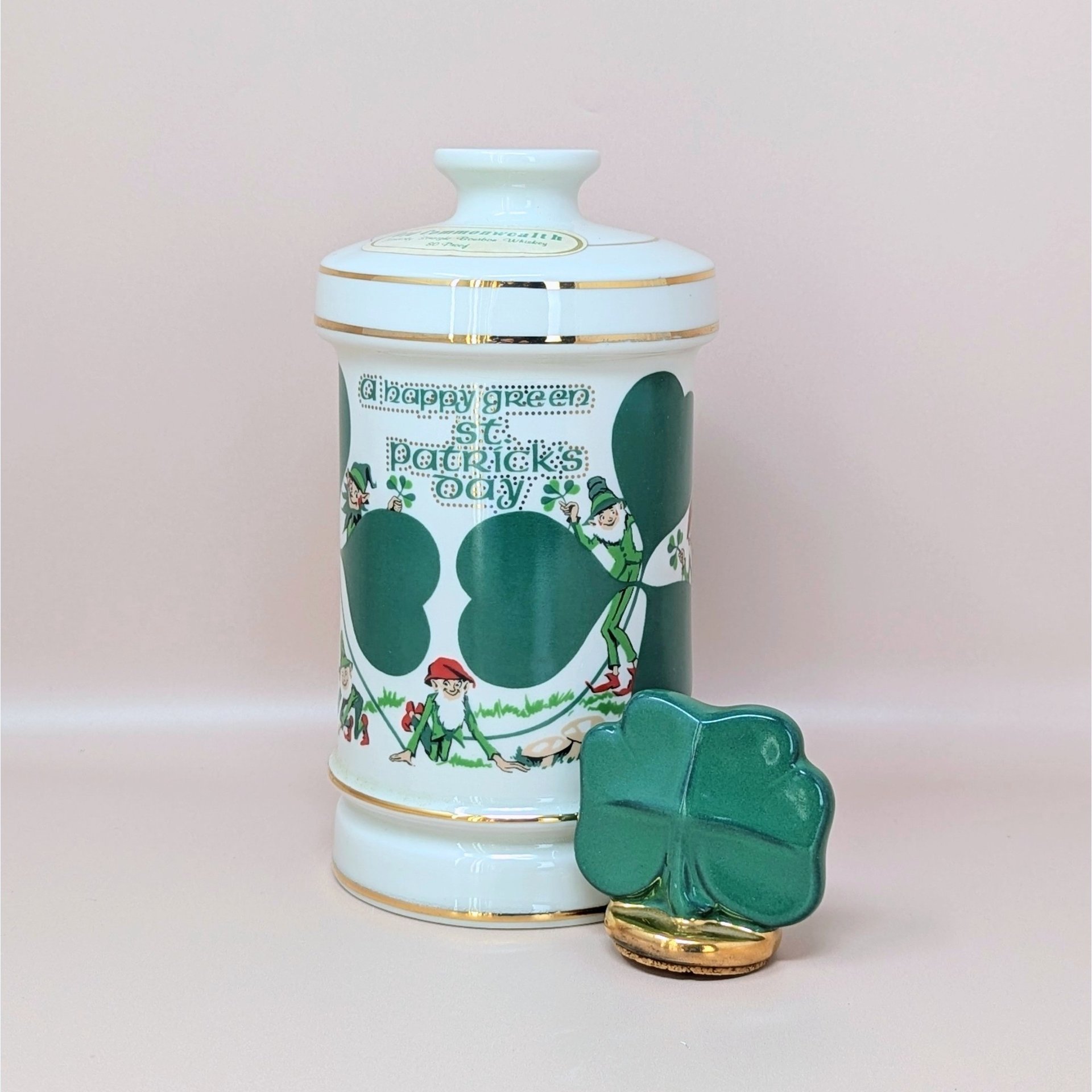 A tall white porcelain decanter featuring green "A Happy Green St. Patrick’s Day" typography, leprechaun illustrations, and a gold-trimmed shamrock stopper against a neutral background.