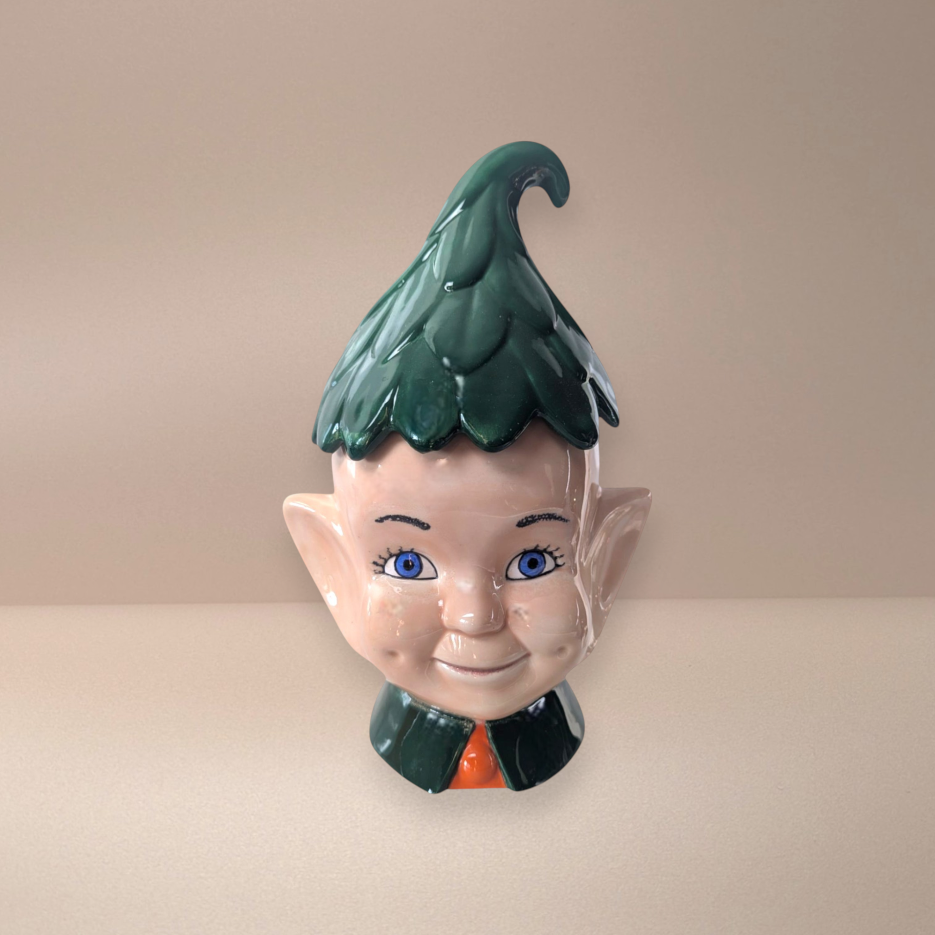 Large ceramic pixie elf cookie jar. Features a smiling face with blue eyes, pointed ears, and a vibrant green leaf-textured hat that serves as the lid. High-gloss glazed finish with deep green and warm flesh tones.