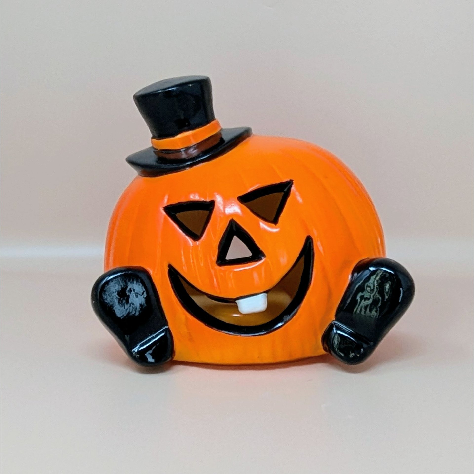 A glossy orange ceramic pumpkin figurine wearing a black top hat and featuring a single white tooth against a neutral background.