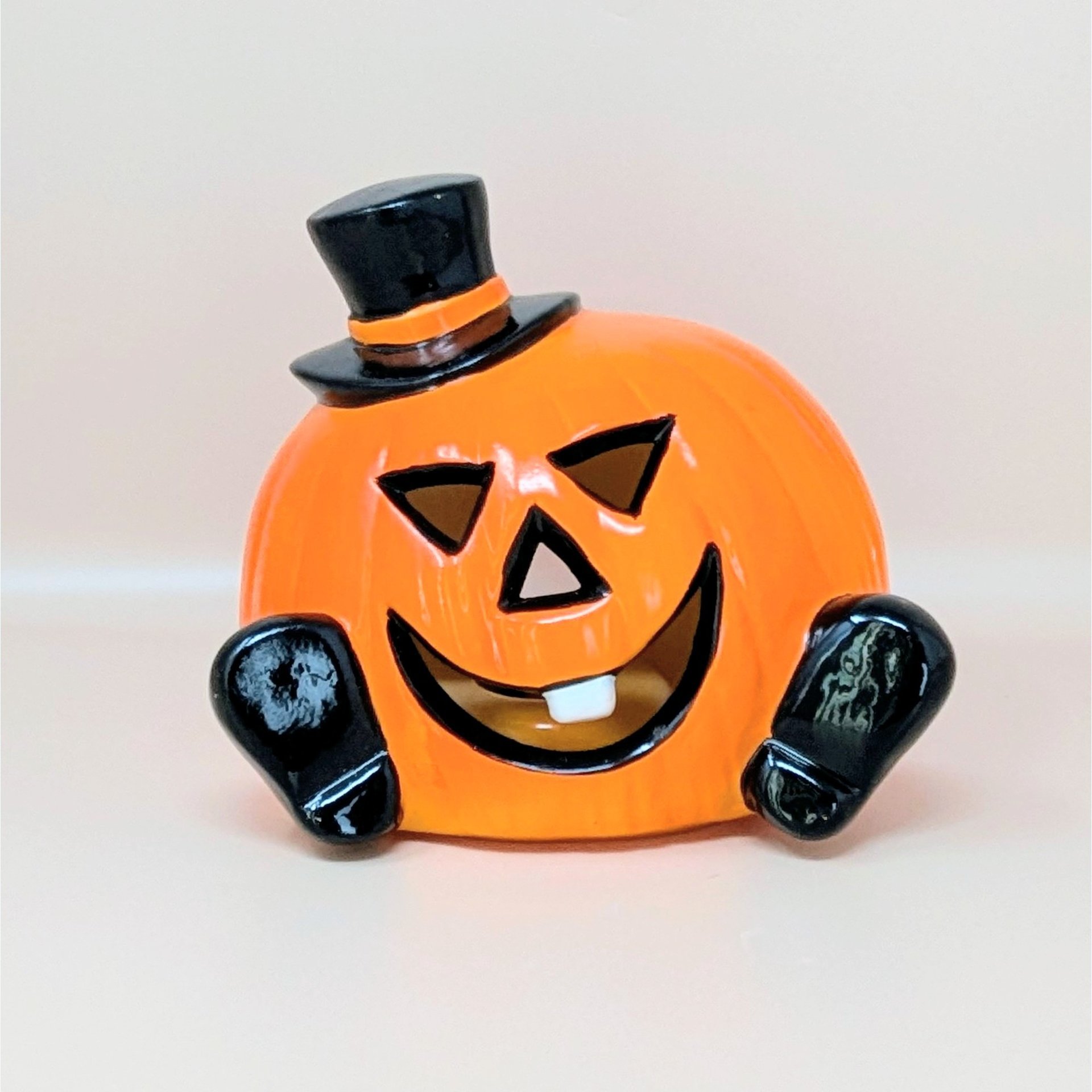 A glossy orange ceramic pumpkin figurine wearing a black top hat and featuring a single white tooth against a neutral background.