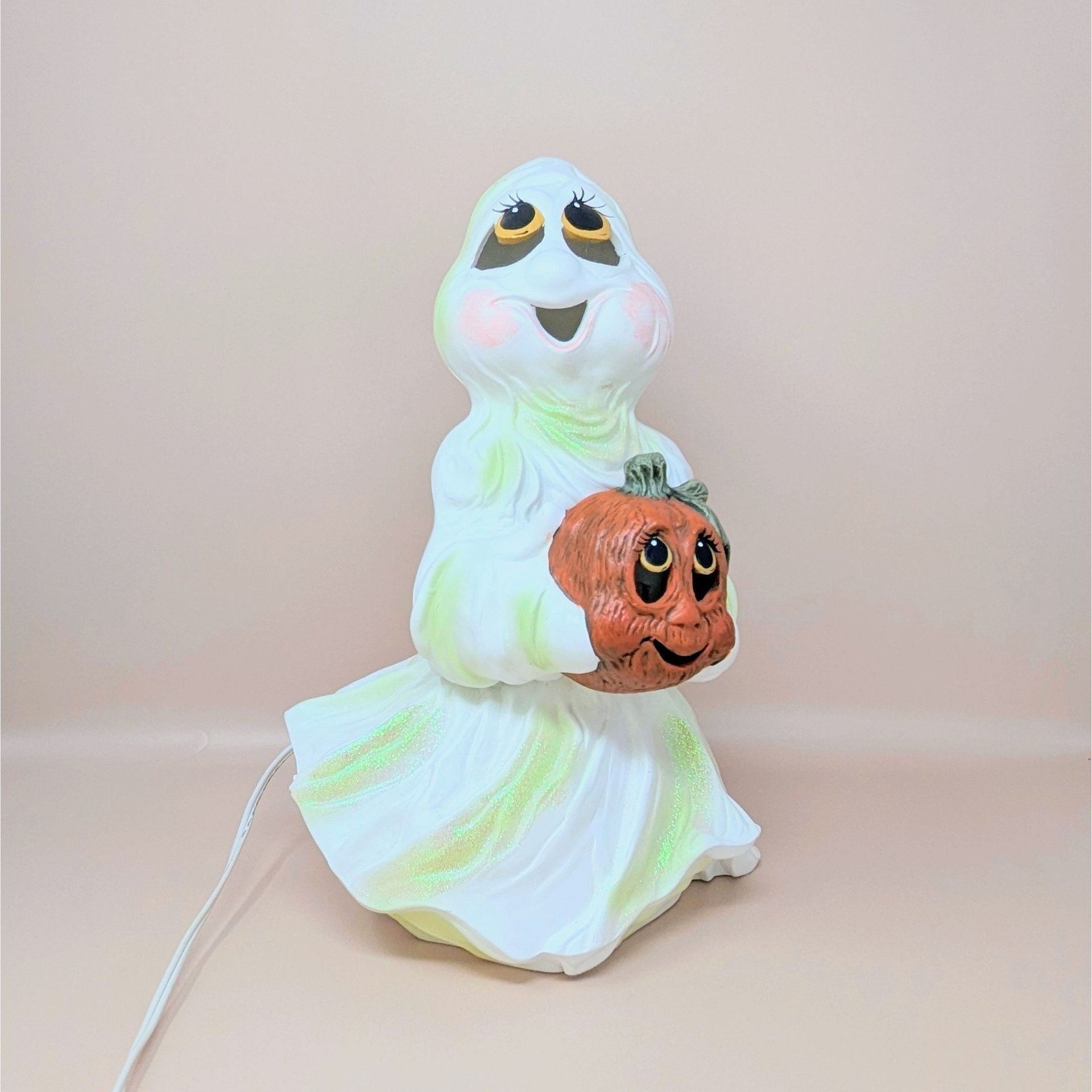A large white ceramic ghost figurine with iridescent green highlights holding an orange jack o' lantern, featuring large expressive eyes and a wide smile.