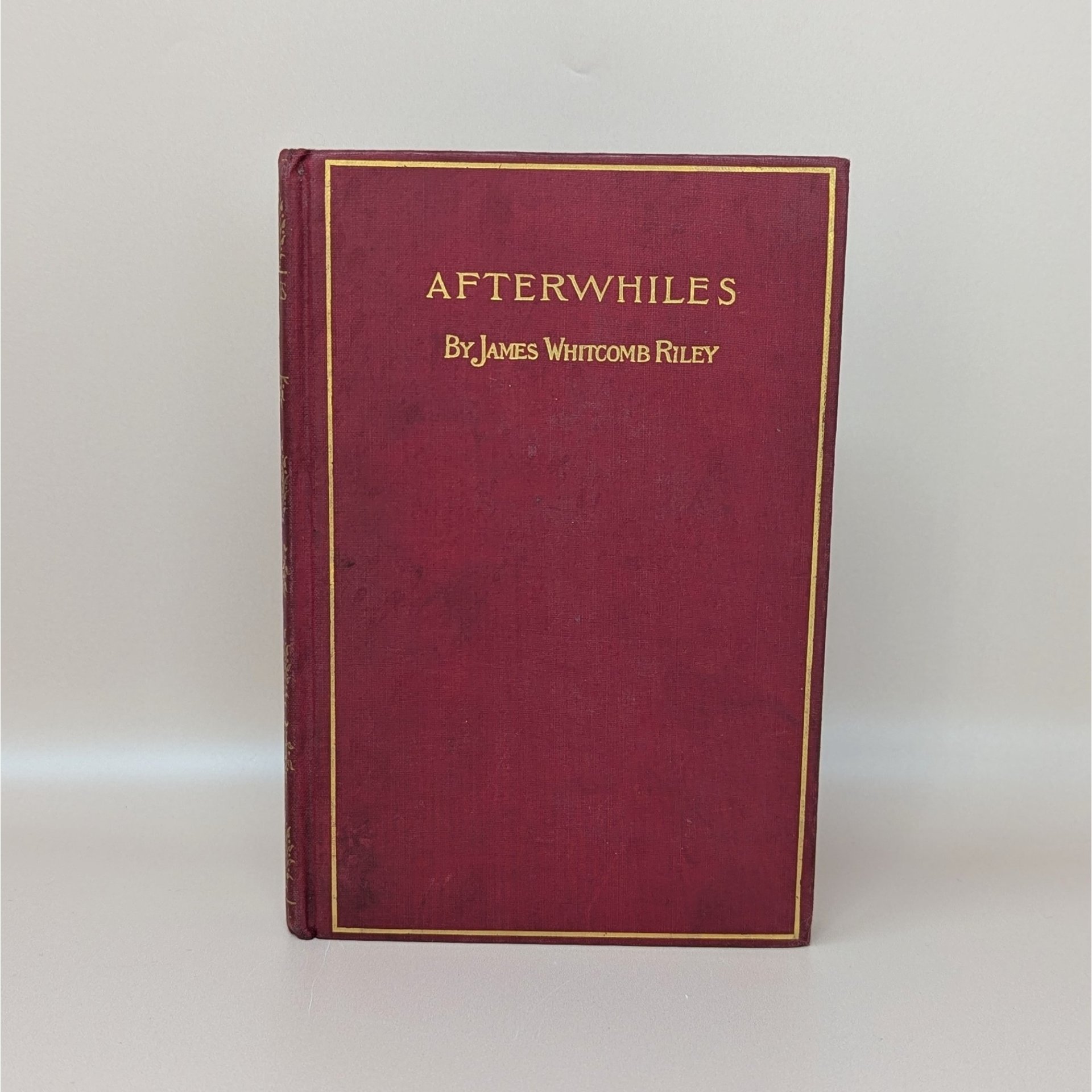A burgundy cloth-bound hardcover book with a gilded rectangular border and gold typography for Afterwhiles by James Whitcomb Riley against a neutral background.