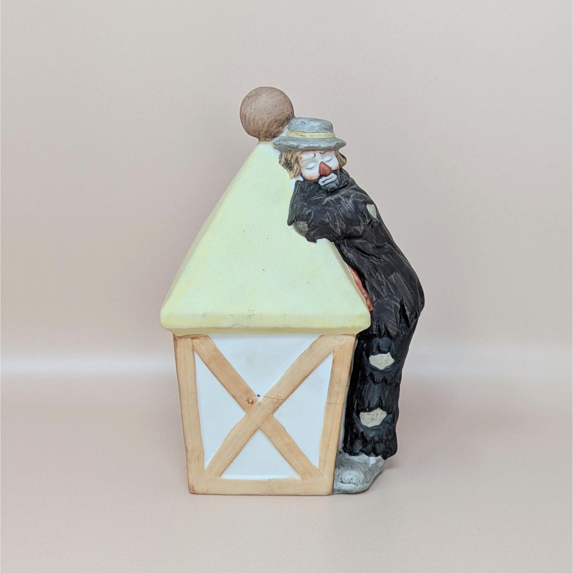 A bisque porcelain figurine of a sad clown in tattered black clothes leaning against a pale yellow street lamp against a neutral background.