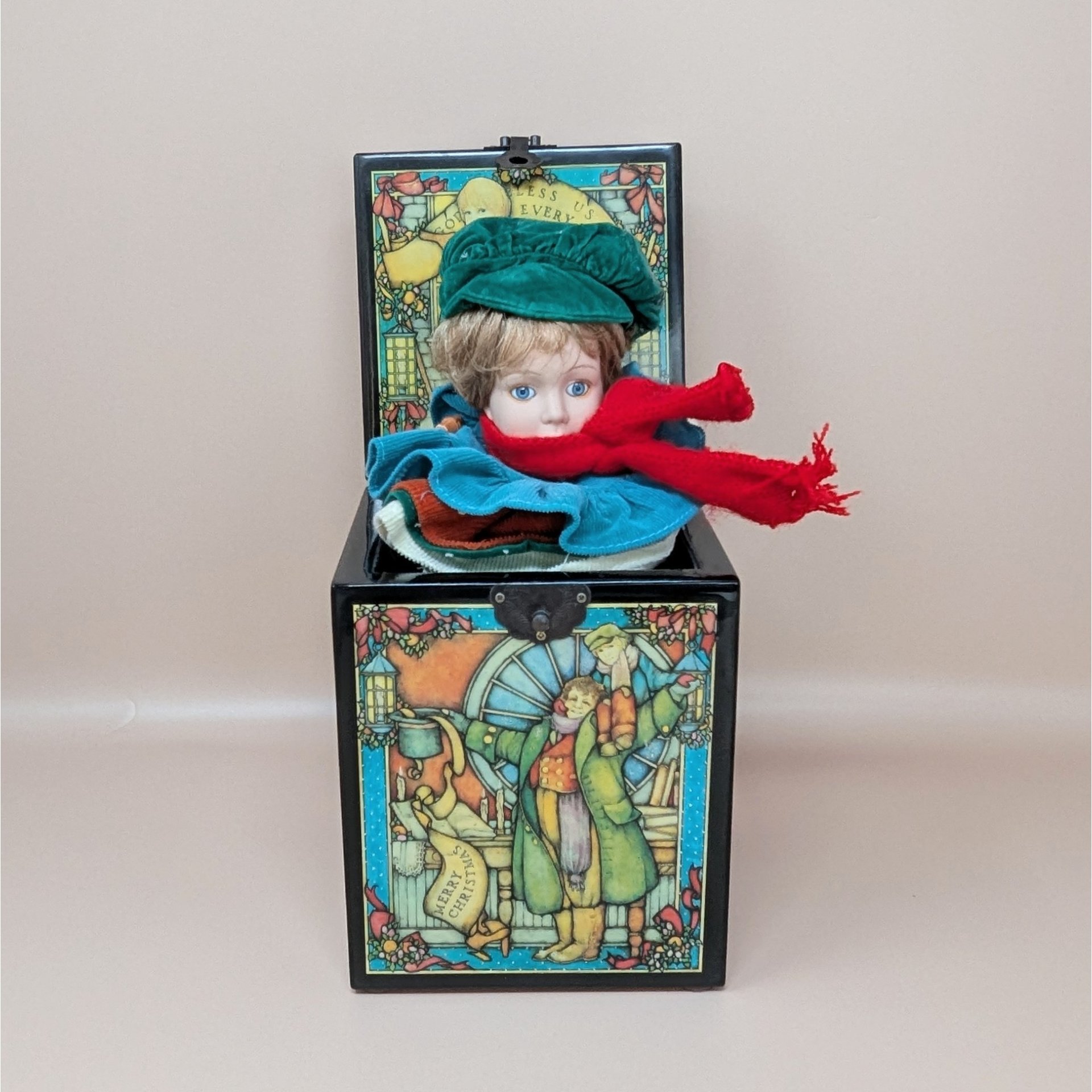 A black wooden music box with an open lid, revealing a porcelain boy doll with a green hat and red scarf. The box features colorful illustrations of Bob Cratchit and Tiny Tim.