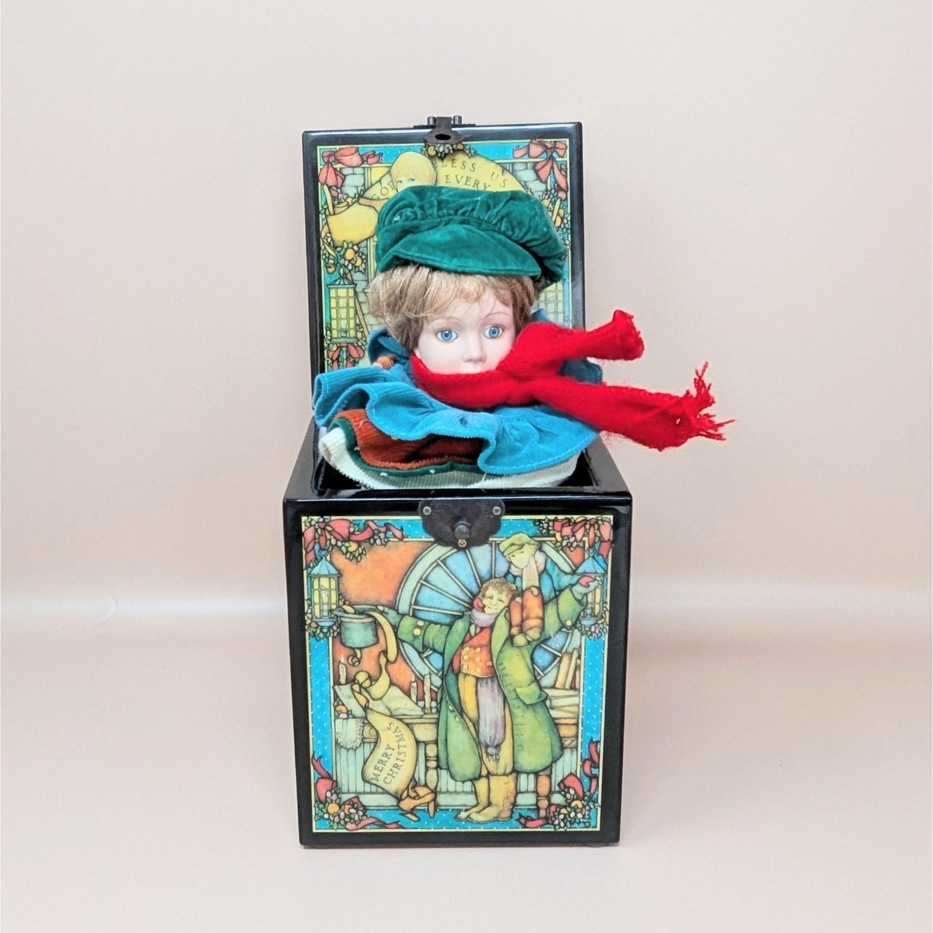 A black wooden music box with an open lid, revealing a porcelain boy doll with a green hat and red scarf. The box features colorful illustrations of Bob Cratchit and Tiny Tim.