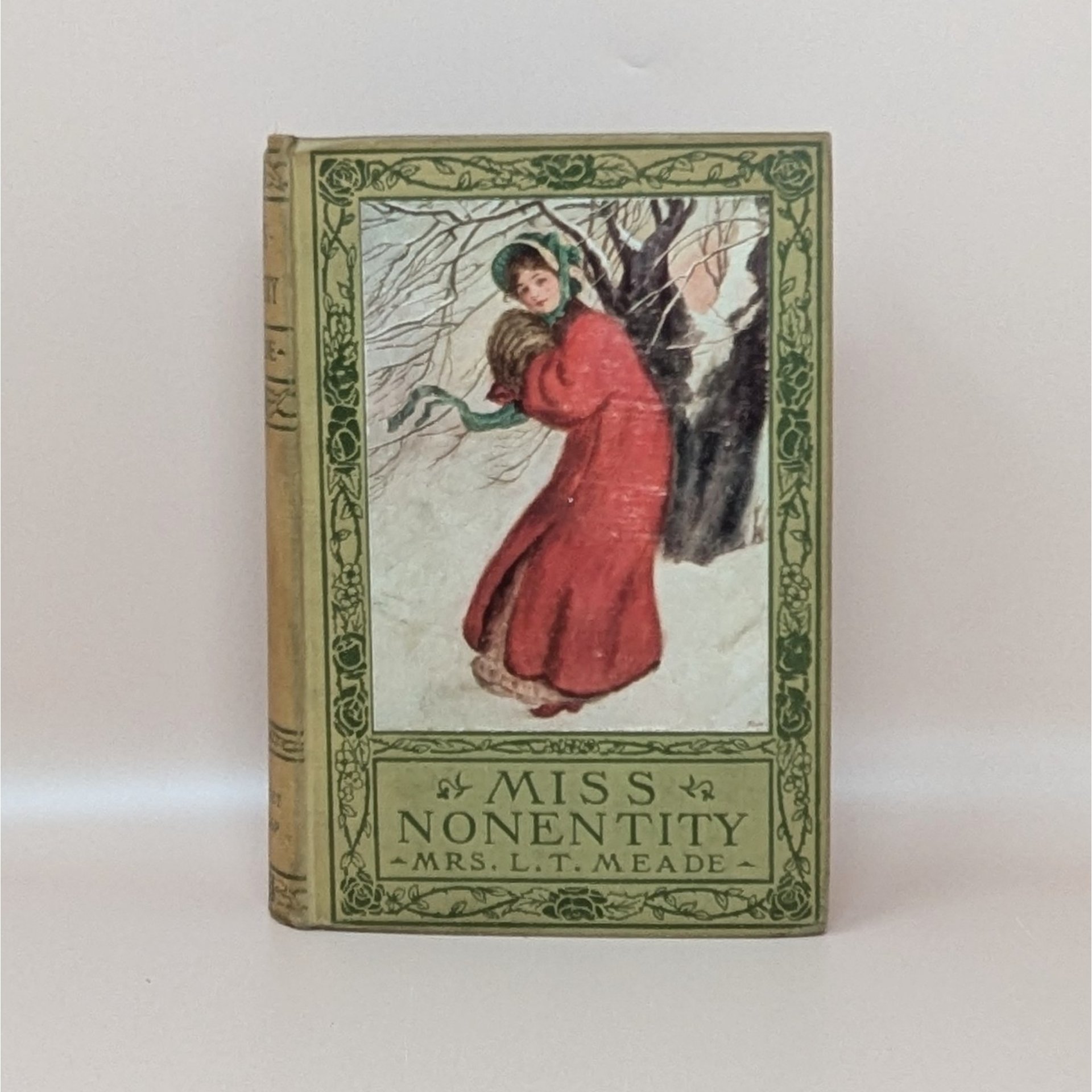 A vintage hardcover book titled Miss Nonentity by Mrs. L.T. Meade, featuring a pictorial cover of a woman in a red winter coat against a snowy background with green floral borders.
