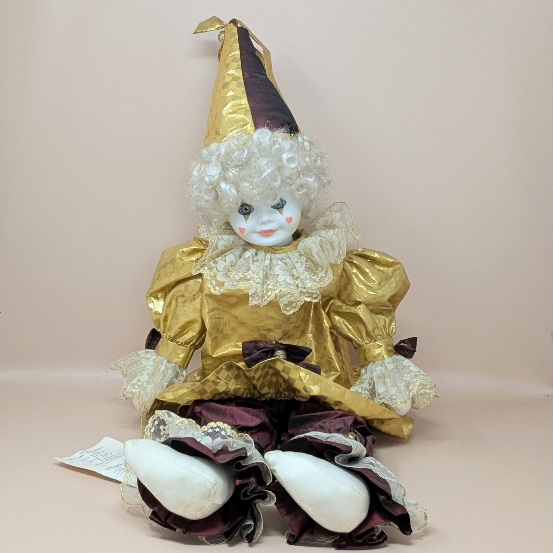 A large porcelain clown doll in a metallic gold and burgundy costume, wearing a tall pointed hat and featuring a white curly wig and hand-painted face.