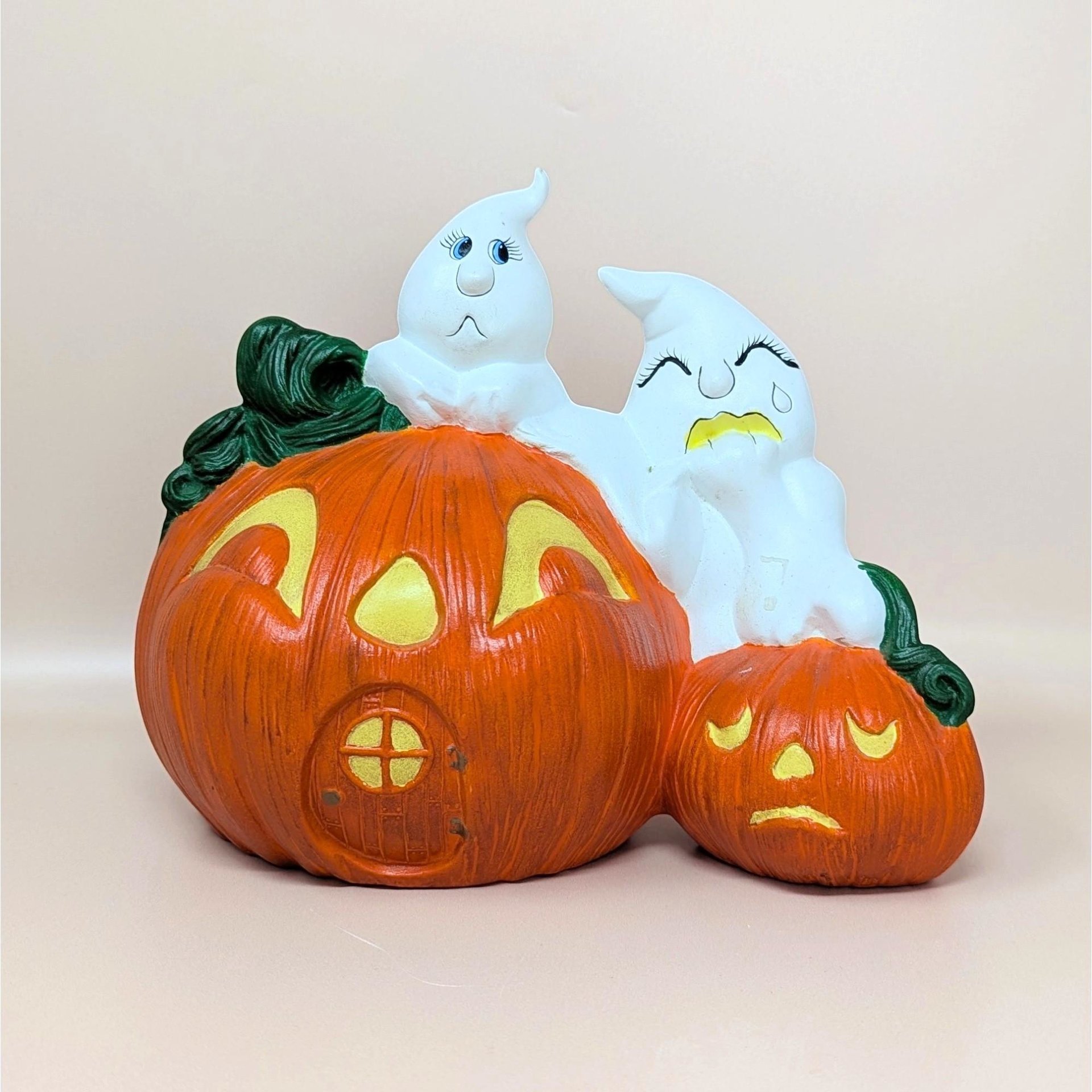 A vintage hand-painted ceramic figurine featuring two white ghosts perched atop a large orange jack o' lantern house and a smaller sad pumpkin.