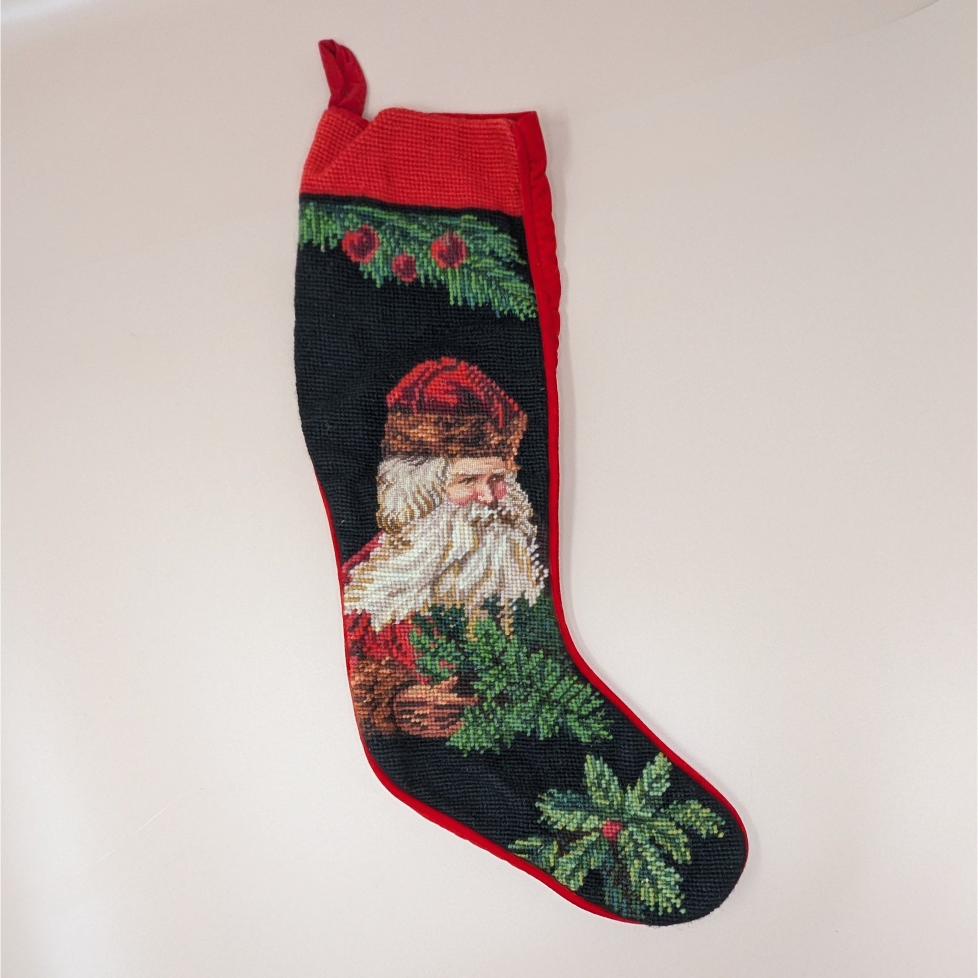 A vintage needlepoint Christmas stocking featuring a detailed Old World Santa with a fur hat and evergreen branches against a dark background.