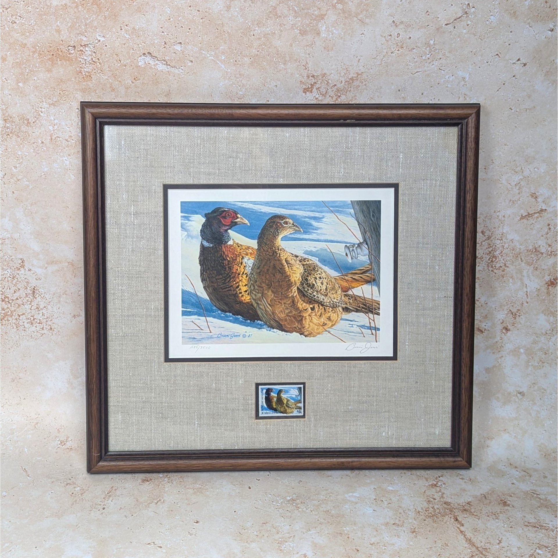 A 1987 Minnesota Pheasant Habitat Stamp Print by Brian Jarvi on a tan textured background. The framed art features a signed and numbered print of a pheasant pair in the snow, a matching stamp below, an oatmeal linen mat, and a dark wood frame.