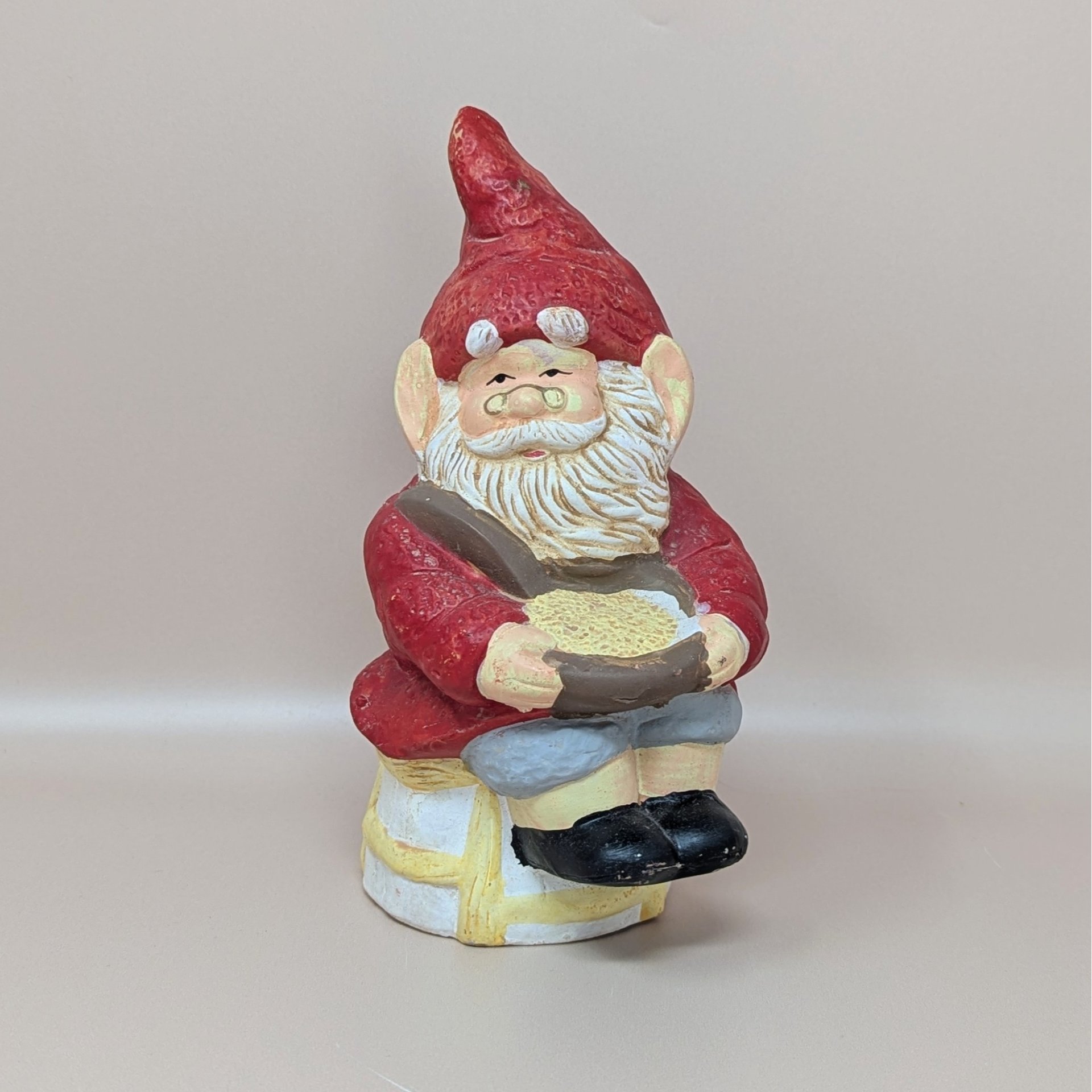 A vintage ceramic gnome figurine with a tall red hat, white beard, and glasses, sitting on a stool and holding a golden bowl.