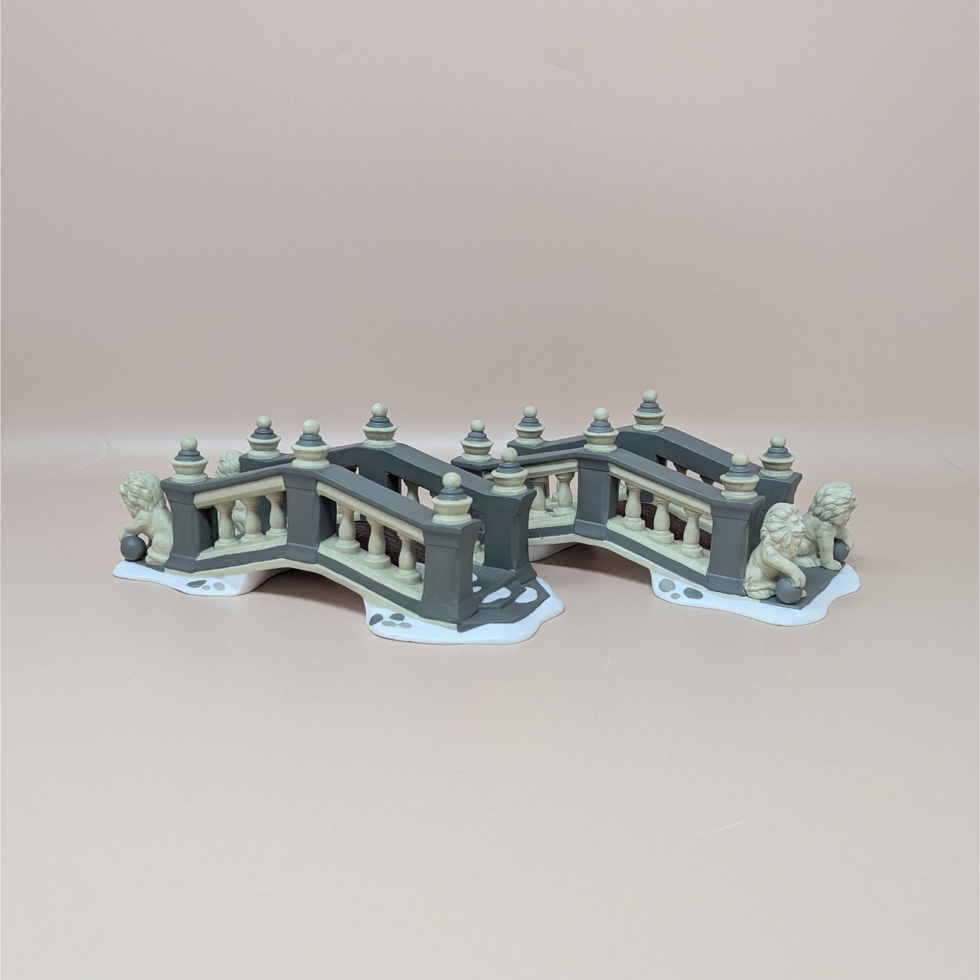 A pair of Department 56 Heritage Village ceramic bridges featuring grey and cream railings with miniature lion statues at the entrance on a snowy base.