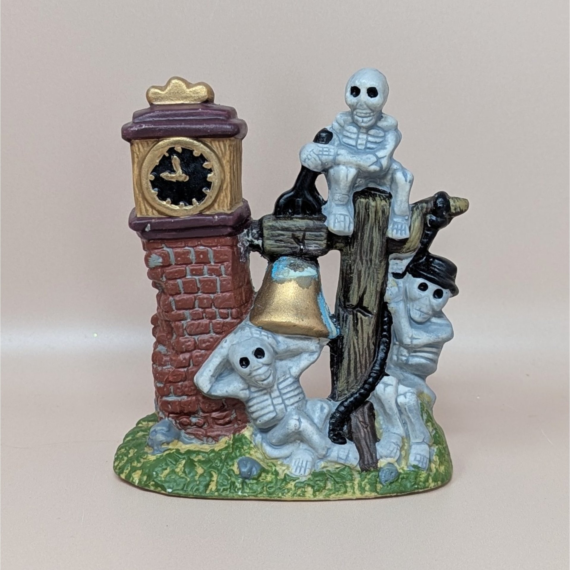 A vintage Halloween village accessory featuring a brick clock tower with three skeletons lounging around a golden bell and a wooden post.