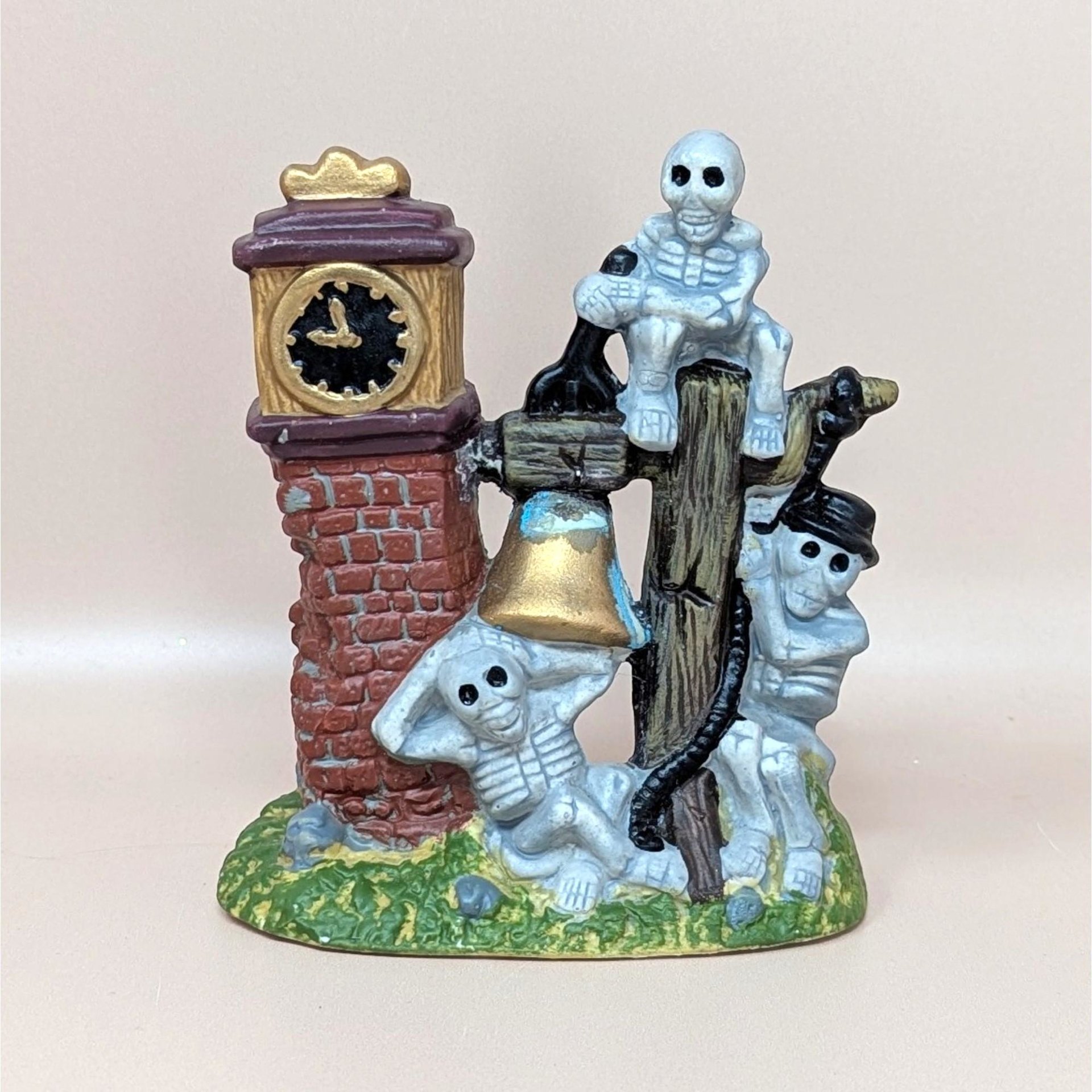A vintage Halloween village accessory featuring a brick clock tower with three skeletons lounging around a golden bell and a wooden post.