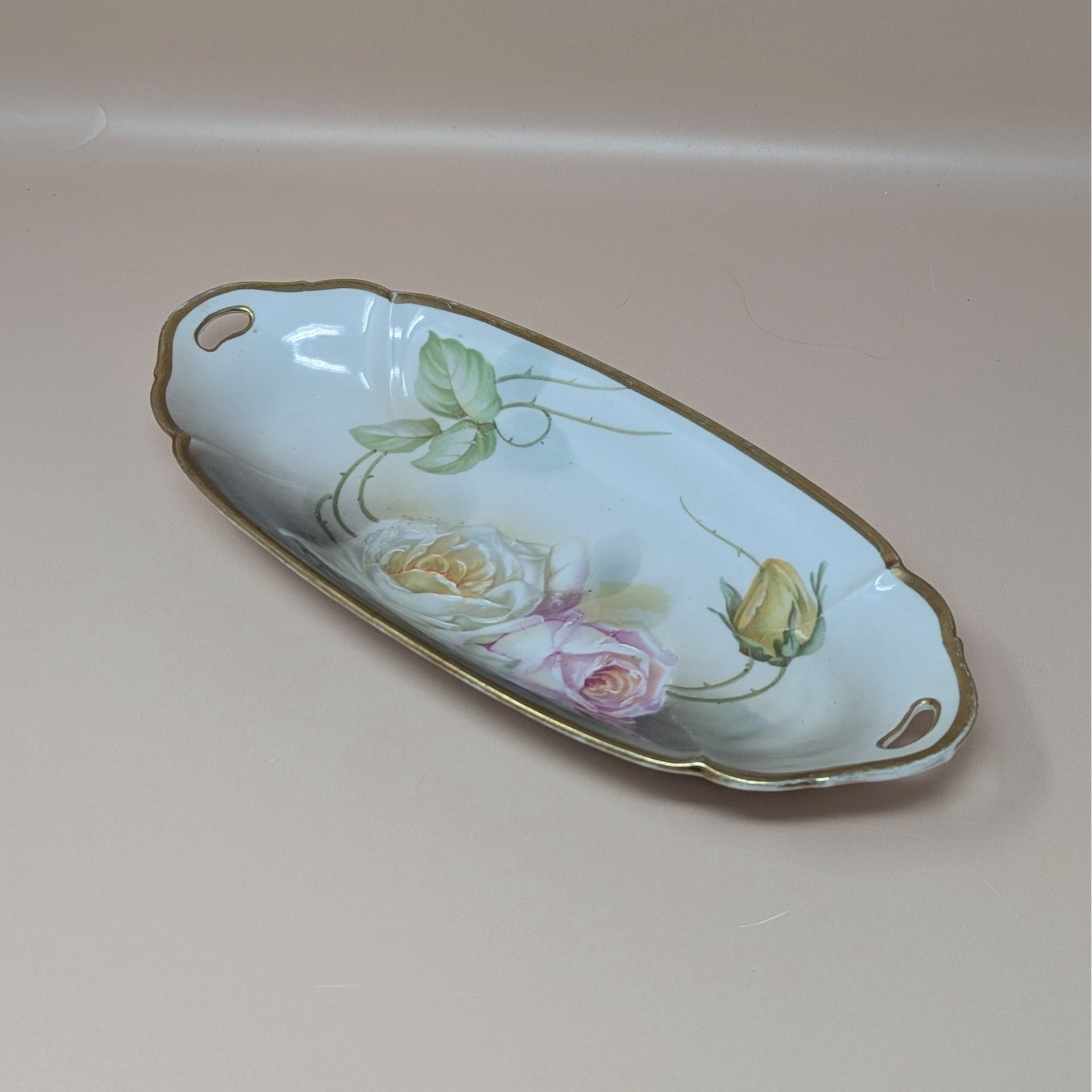 A vintage porcelain celery tray from Suhl, Germany, featuring a long oval shape with hand-painted yellow and pink roses and a worn gold rim.