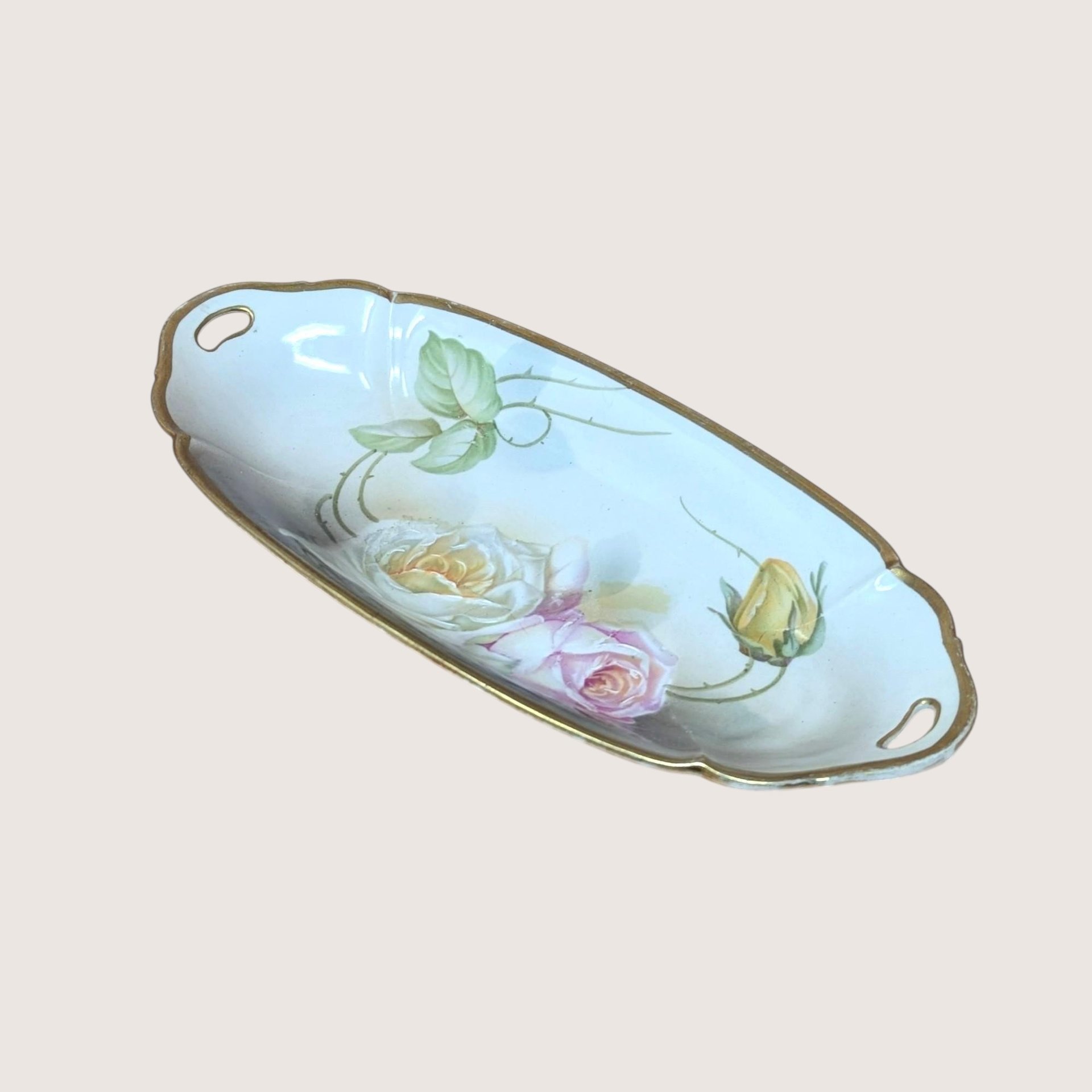 An oval porcelain tray with painted roses and gold trim, angled on a beige background.