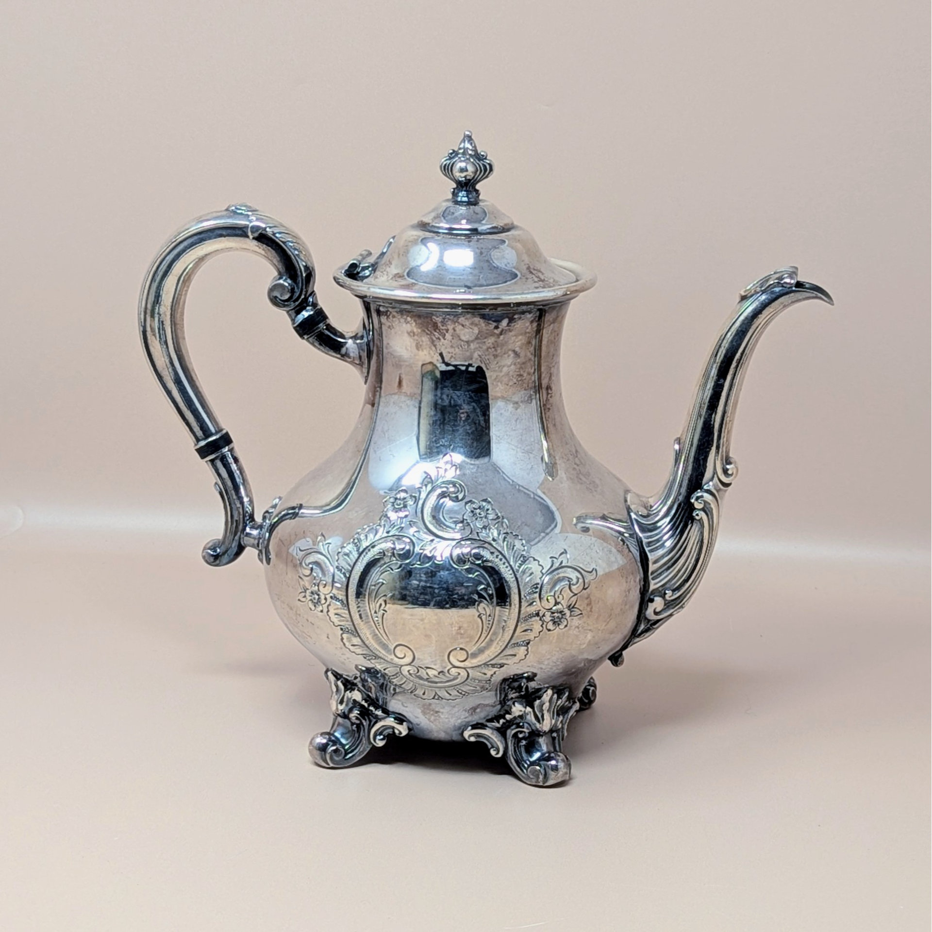 A vintage silver plated teapot with ornate hand chased floral scrollwork and a decorative finial on the lid, standing on four scrolled feet