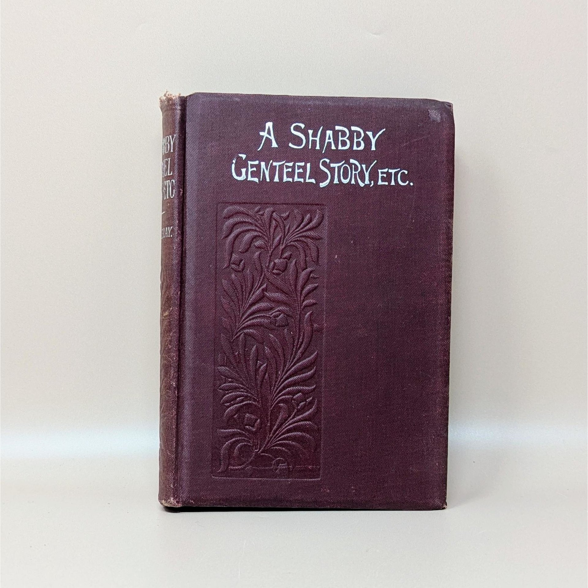 An antique burgundy hardcover book titled A Shabby Genteel Story by Thackeray with an embossed floral panel and silver-toned lettering