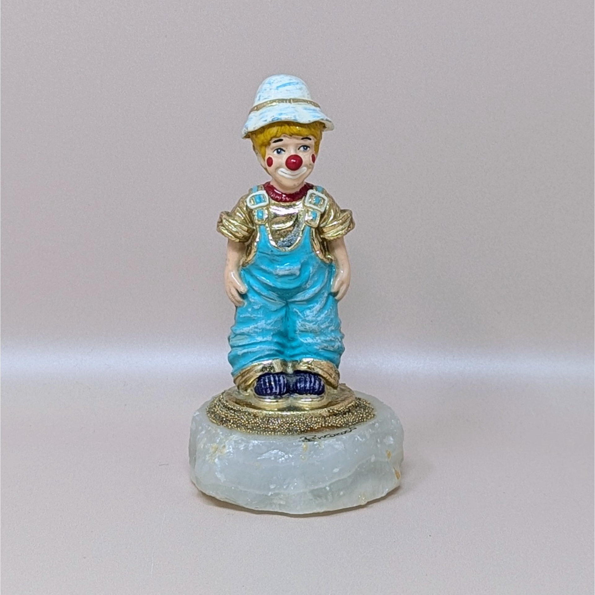 A vintage hand painted clown figurine by Ron Lee named Rascal, wearing blue overalls and a white hat, mounted on a thick natural onyx stone base