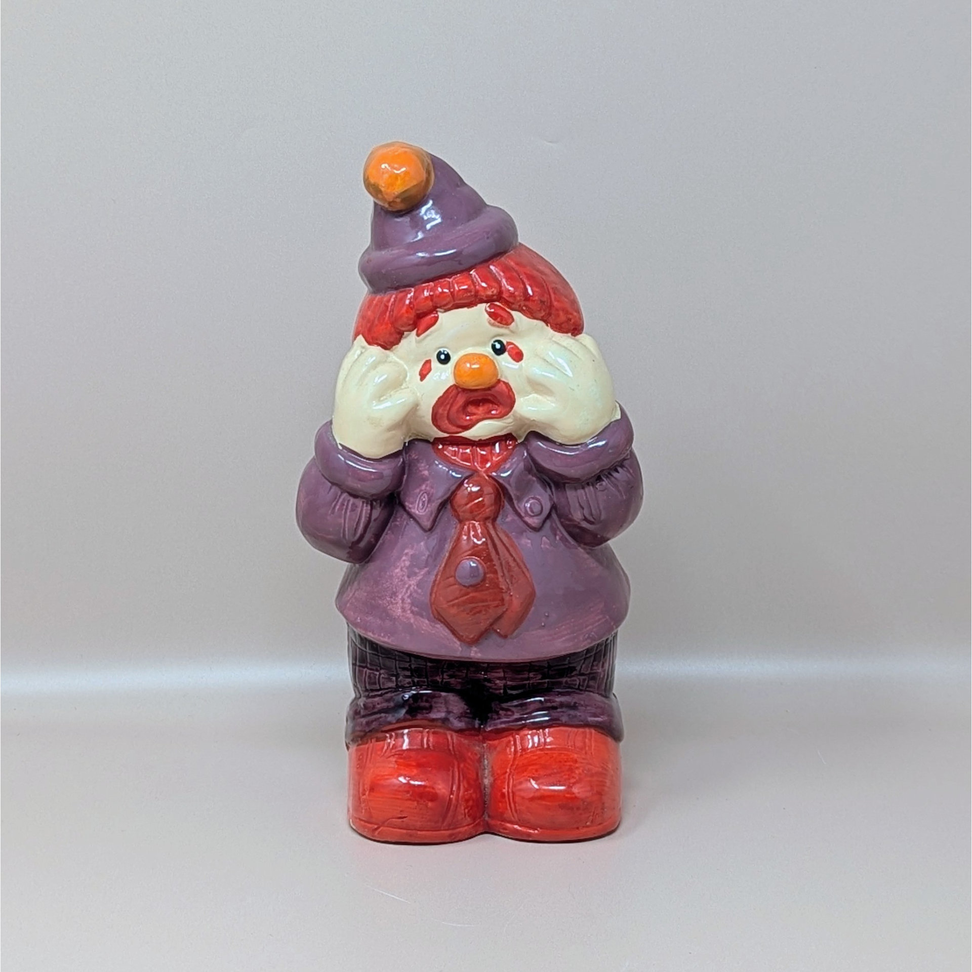 A vintage handpainted ceramic figurine of a crying clown wearing a purple suit and red tie with his hands to his face