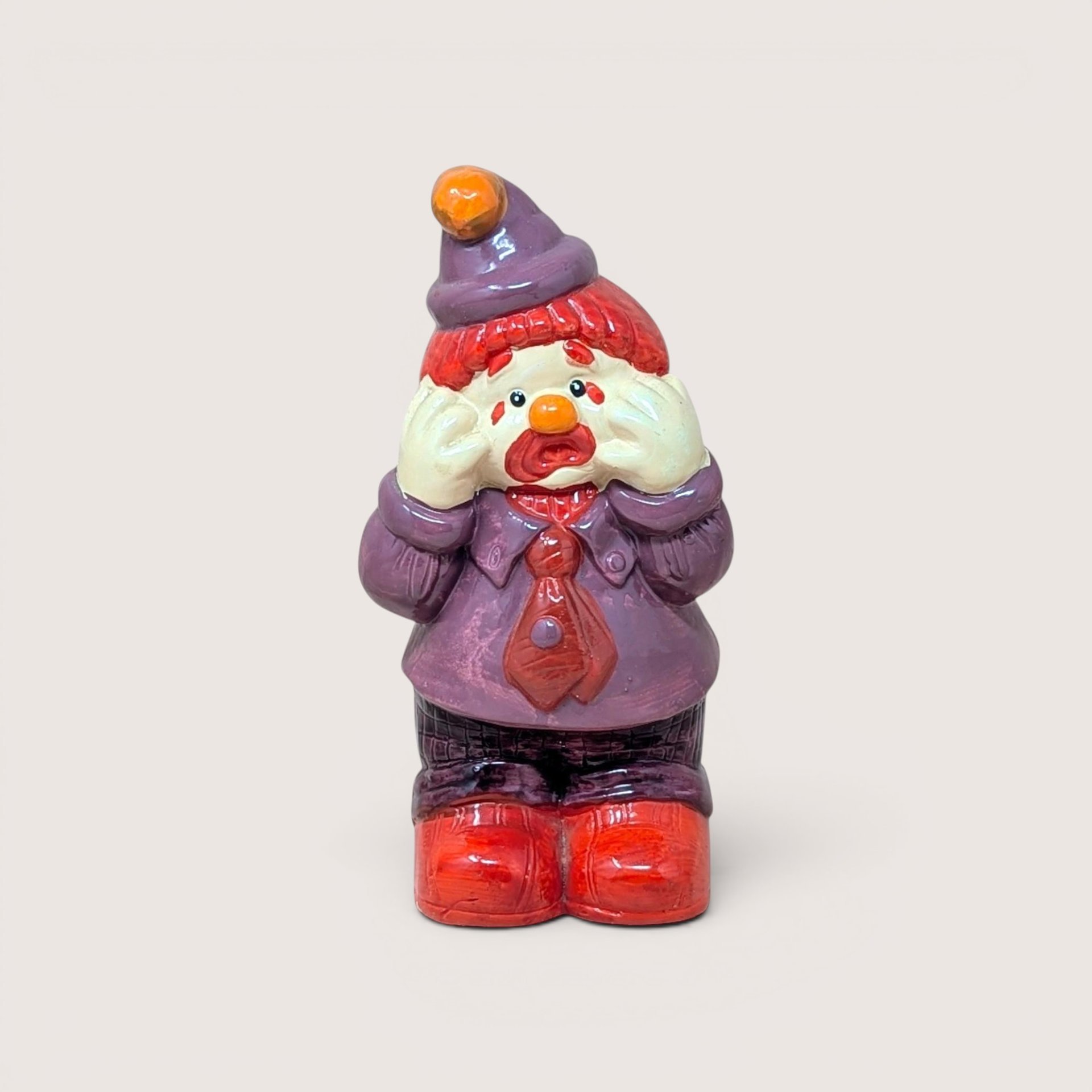 A ceramic sad clown figurine wearing a purple suit and hat with its hands on its face, on a beige background.