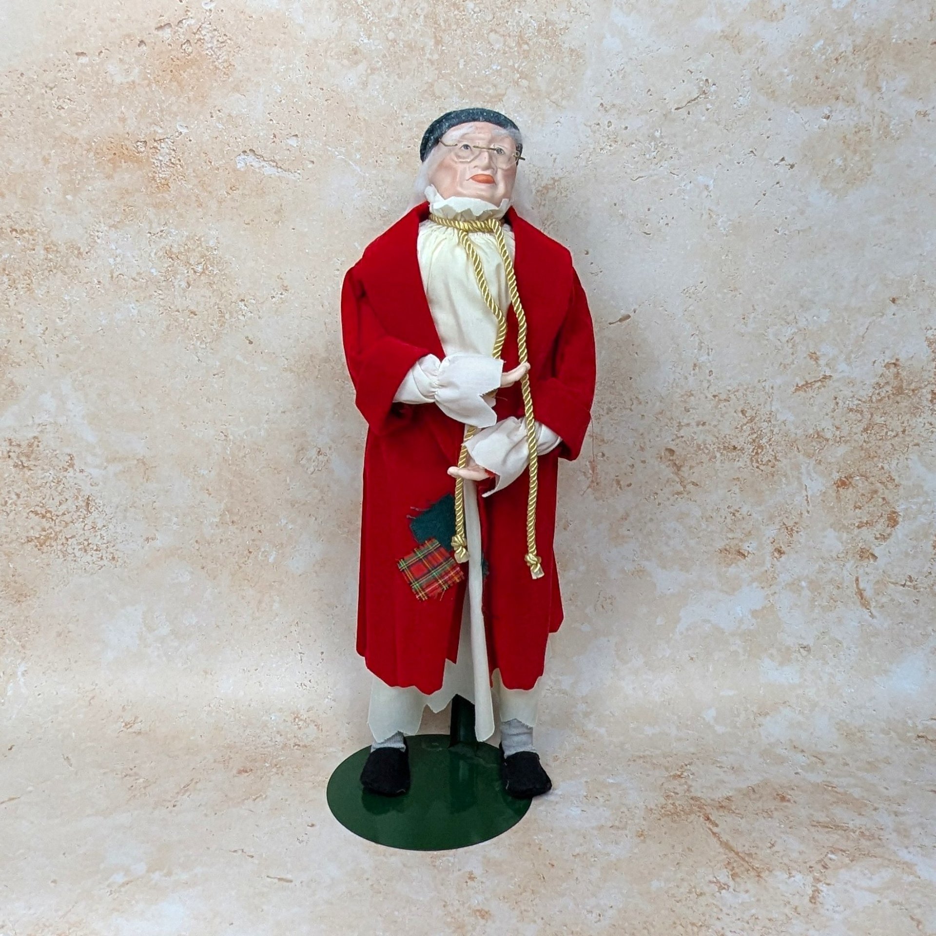 A 1987 vintage Department 56 Ebenezer Scrooge doll on a tan textured background. The nineteen inch tall figure features a detailed porcelain face with wire glasses, dressed in a red velvet night robe and holding a gold rope cord on a green metal stand.